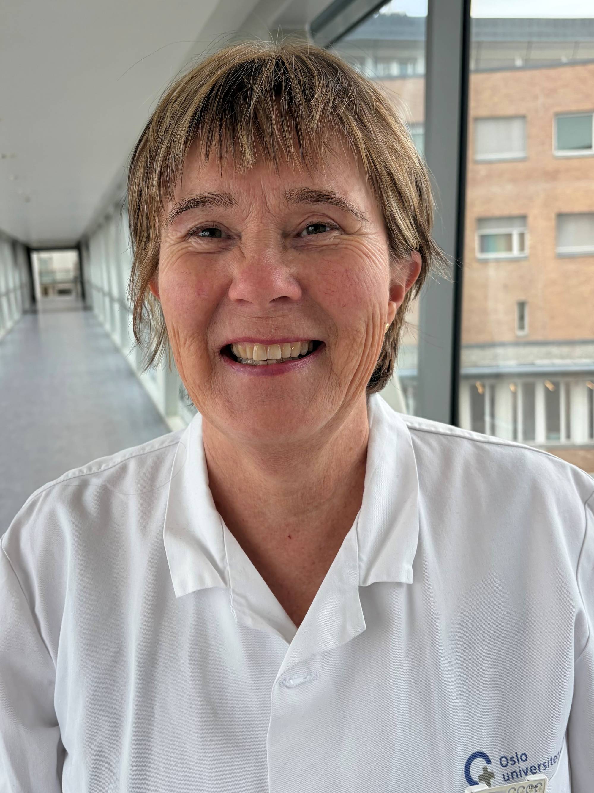 Irene Andersen står i en sykehuskorridor, iført hvite arbeidsklær