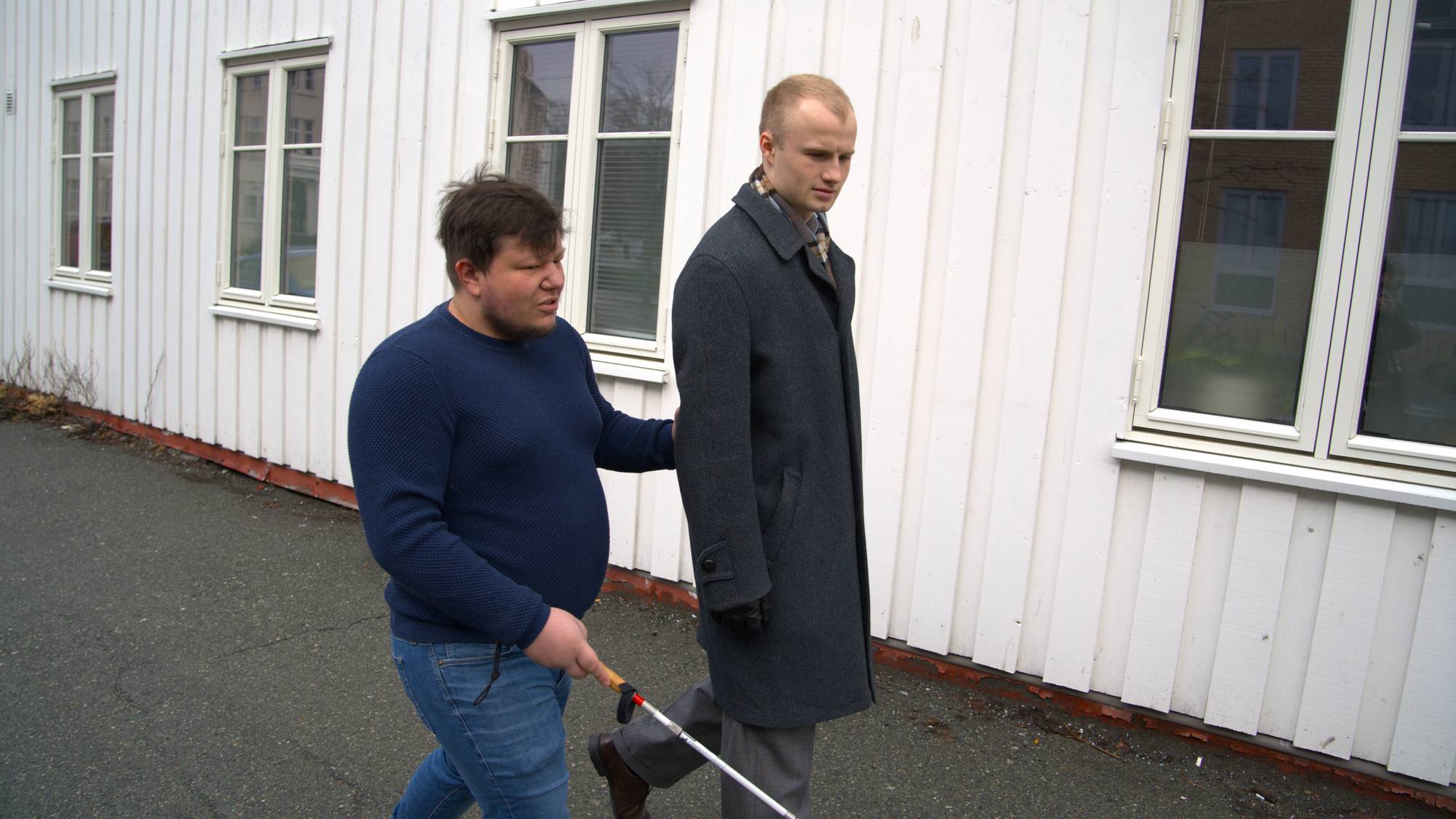 Bildet viser en blind man som går sammen med sin støttekontakt. De går på fortauet langs veggen til et hus i bakgrunn.