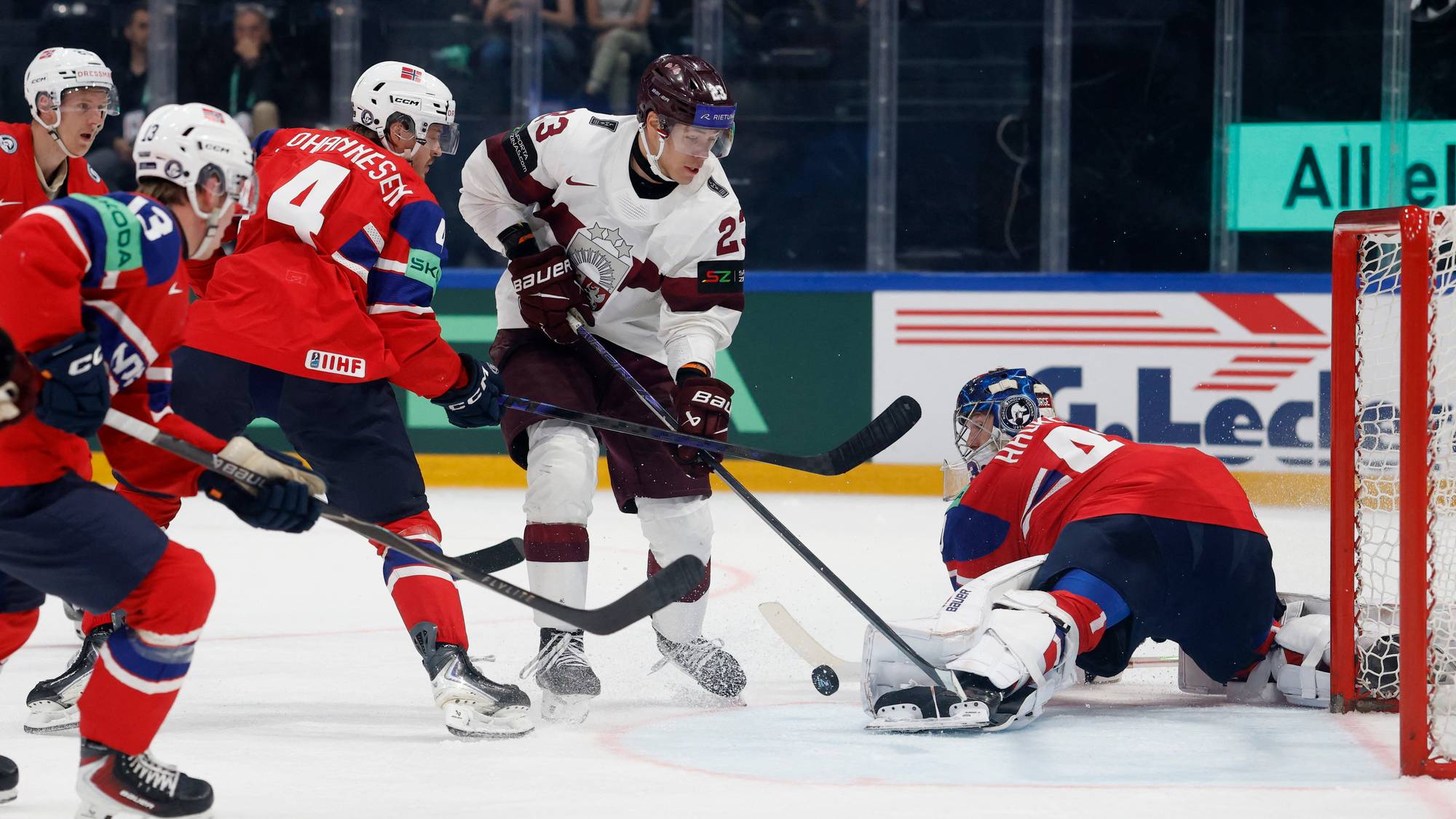 Ishockeyspiller i hvit drakt med nummer 21, og spillere i rød drakt. En målvakt er i knehøy stilling foran et målnett. Det er flere spillere som deltar i kampen på isen. (Bildebeskrivelsen er laget av en KI-tjeneste)
