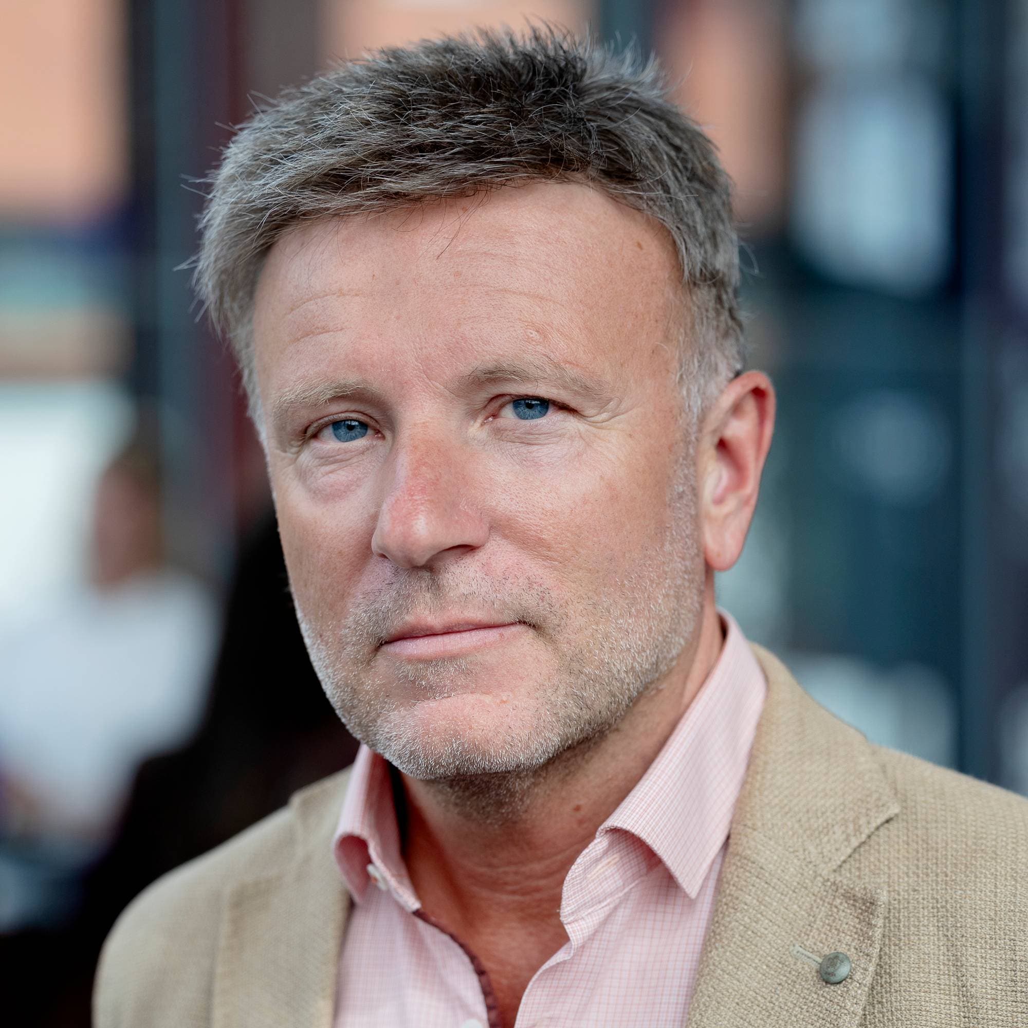 Mann med gråsprengt hår og blå øyne. Han har på seg en lys rosa skjorte og en beige blazer. Bakgrunnen er uskarp og viser bygninger. (Bildebeskrivelsen er laget av en KI-tjeneste)