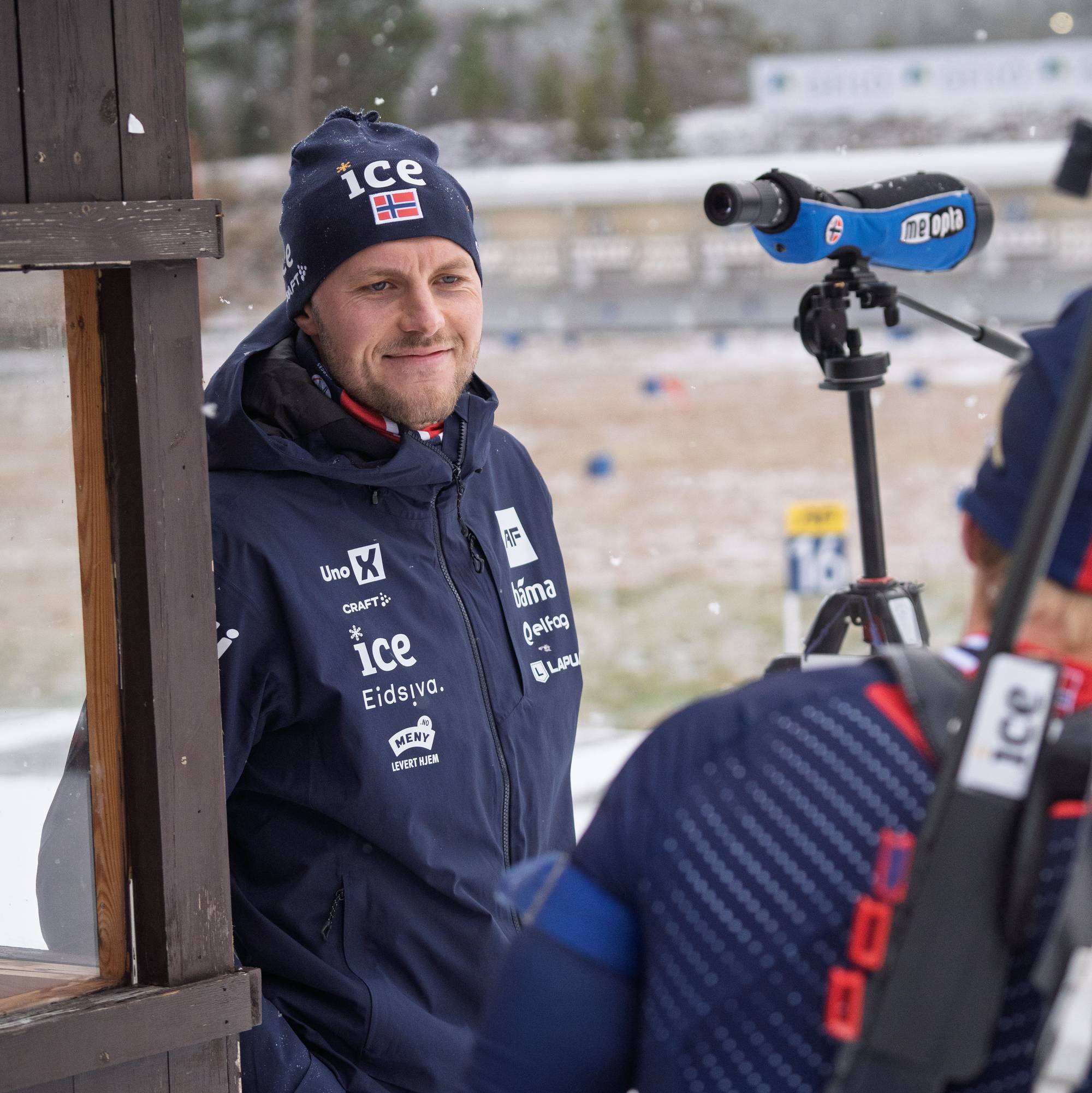To personer står i en skytterbås under snøvær. Den ene er kledd i en mørk jakke med logoer fra sponsorer. Den andre personen har på seg en rød og blå jakke med det norske flagget og holder et skytesiktemiddel. I bakgrunnen er det flere skiver og en snødekt skisportsarena. (Bildebeskrivelsen er laget av en KI-tjeneste)