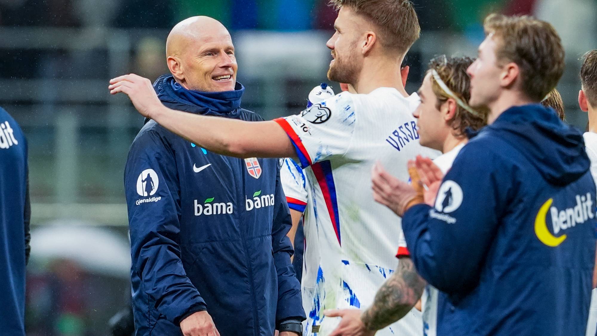 Bildet viser flere mennesker på en fotballbane, sannsynligvis etter en kamp. En spiller i hvit drakt omfavner en annen, mens flere personer i treningsklær applauderer i bakgrunnen. Det er regn i luften, og omgivelsene ser ut til å være en stadion med grønne seter. Uttrykkene på ansiktene indikerer en åpenbar følelse av samhold og glede. (Bildebeskrivelsen er laget av en KI-tjeneste)