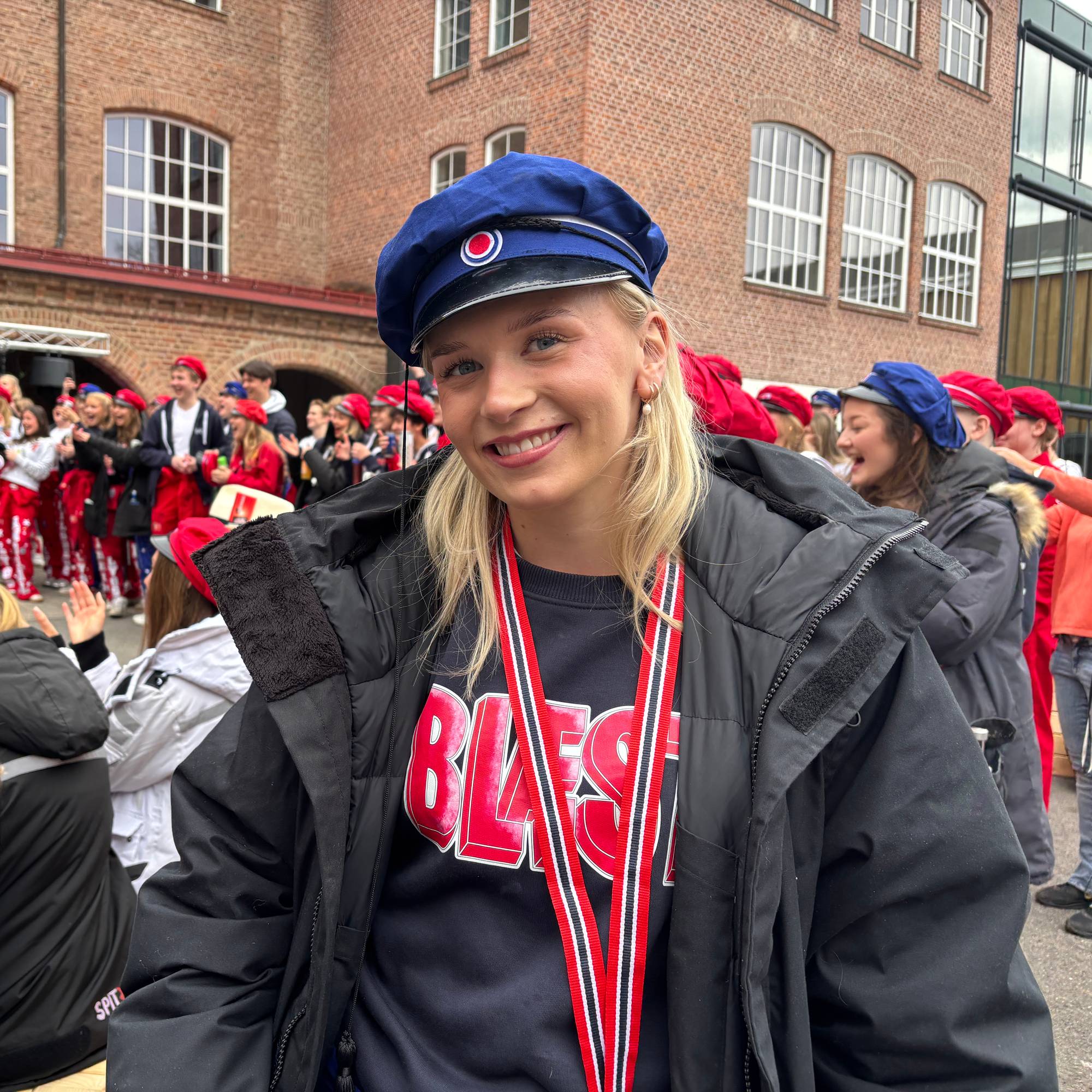 En ung kvinne med blå hatt og russekjole ser mot kamera. Bak henne står flere personer kledd i røde og hvite russekostymer. 