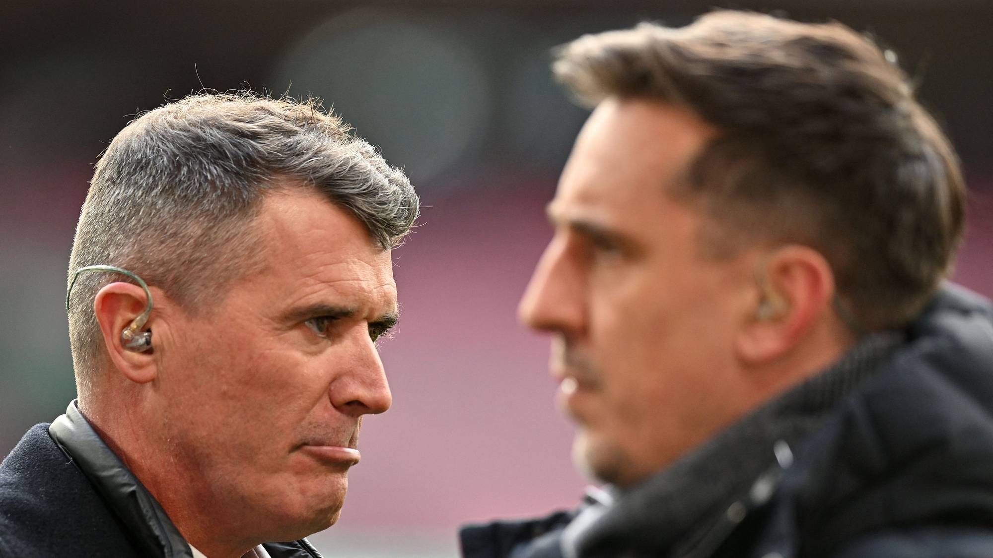 To fotballkommentatorer, Roy Keane og Gary Neville, ser på hverandre. Begge mennene har på seg ørepropper for kommunikasjon. Bakgrunnen er uskarpt og viser muligens et stadion eller et annet sportsarrangement. (Bildebeskrivelsen er laget av en KI-tjeneste)