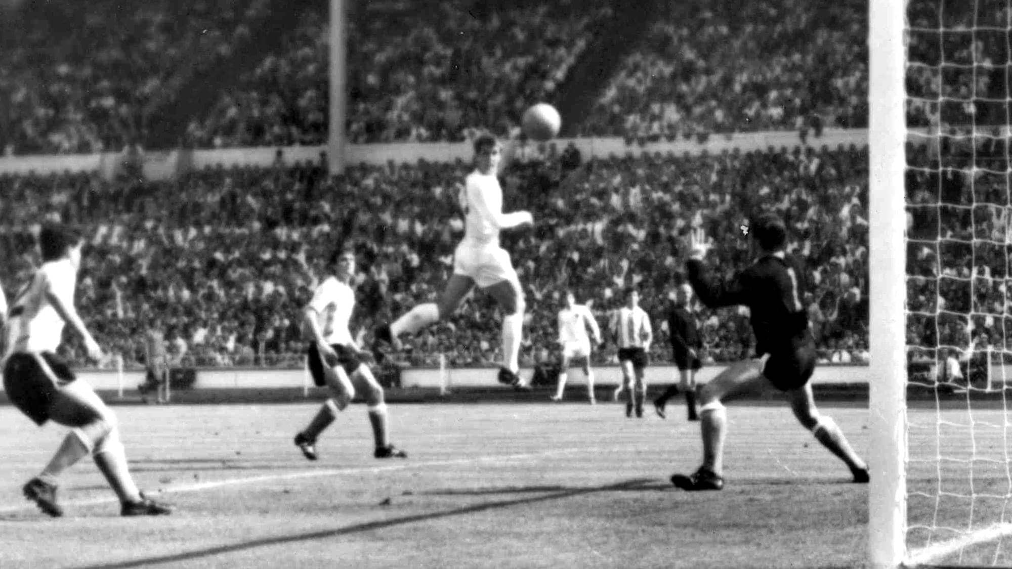Fotballspiller i svev mot mål foran keeper under en kamp på en fullsatt stadion. Svart-hvitt foto. (Bildebeskrivelsen er laget av en KI-tjeneste)