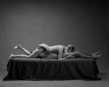 Et nakent par ligger med hodet mot hverandres kjønnsorgan på en svart seng. Mannen har mørkt hår og tatoveringer. Kvinnen har blondt hår og ligger øverst. Mannen holder hendene på rumpeballene til kvinnen. De simulerer oral sex.