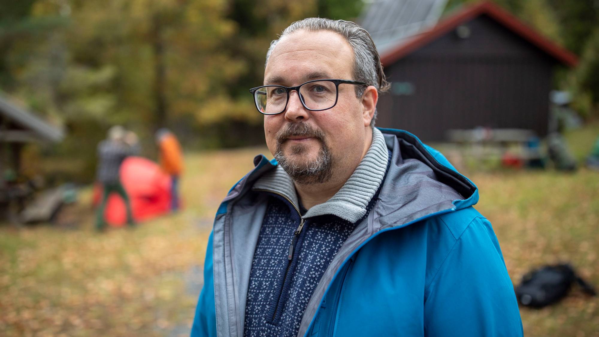 Portrett av en mann i blå jakke utendørs ved en hytte, med personer i bakgrunnen. (Bildebeskrivelsen er laget av en KI-tjeneste)