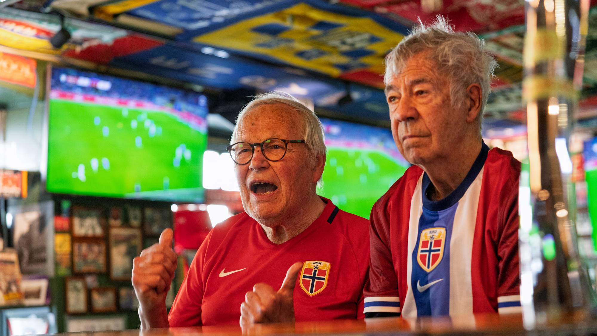 Arne Scheie og Egil Olsen sitter ved et bord på en pub. Begge er iført fotballdrakter med det norske flagget. I bakgrunnen henger det mange fotballskjerf og bilder på veggene. En TV-skjerm viser en fotballkamp. 