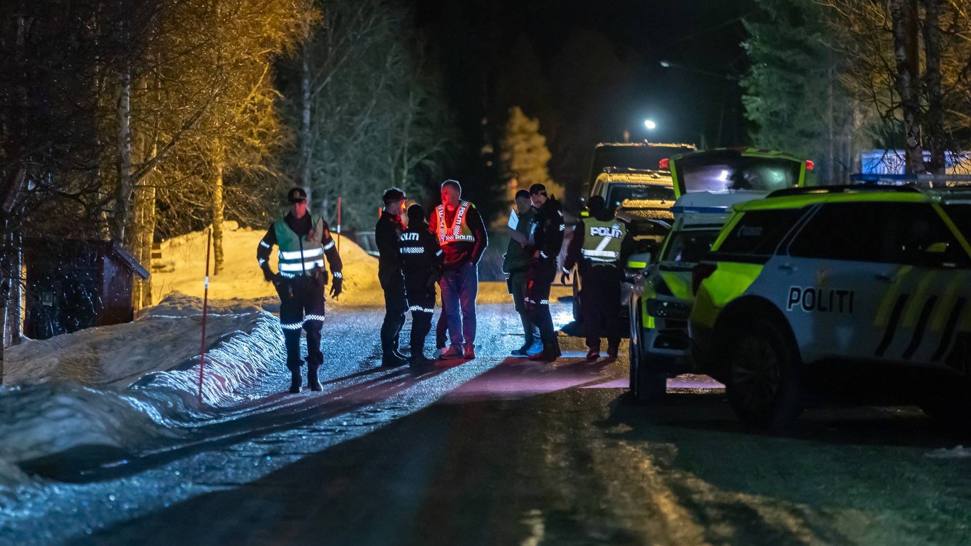 Politimenn og annen nødhjelpspersonell er samlet på en snødekt vei om natten. De er involvert i en samtale, med flere politibiler parkert i nærheten. Omgivelsene er preget av trær og snø, med lys fra bilene som skaper en dramatisk stemning. Det ser ut til å være en aktiv hendelse som krever deres oppmerksomhet. (Bildebeskrivelsen er laget av en KI-tjeneste)