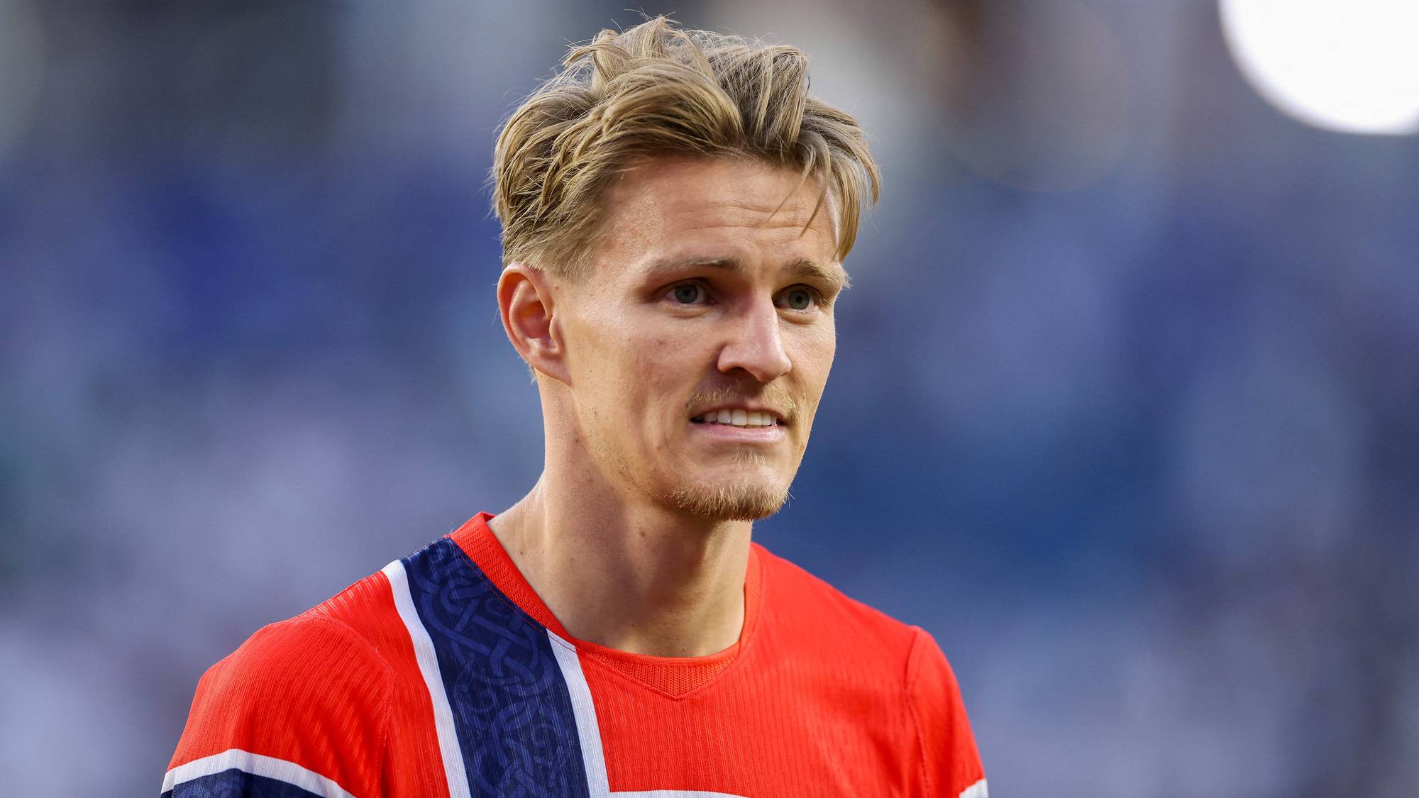 Norsk fotballspiller Martin Ødegaard ser til høyre for kameraet. Han har på seg en rød landslagsdrakt. Bak ham er det uskarpe blå og hvite farger fra et stadion. (Bildebeskrivelsen er laget av en KI-tjeneste)
