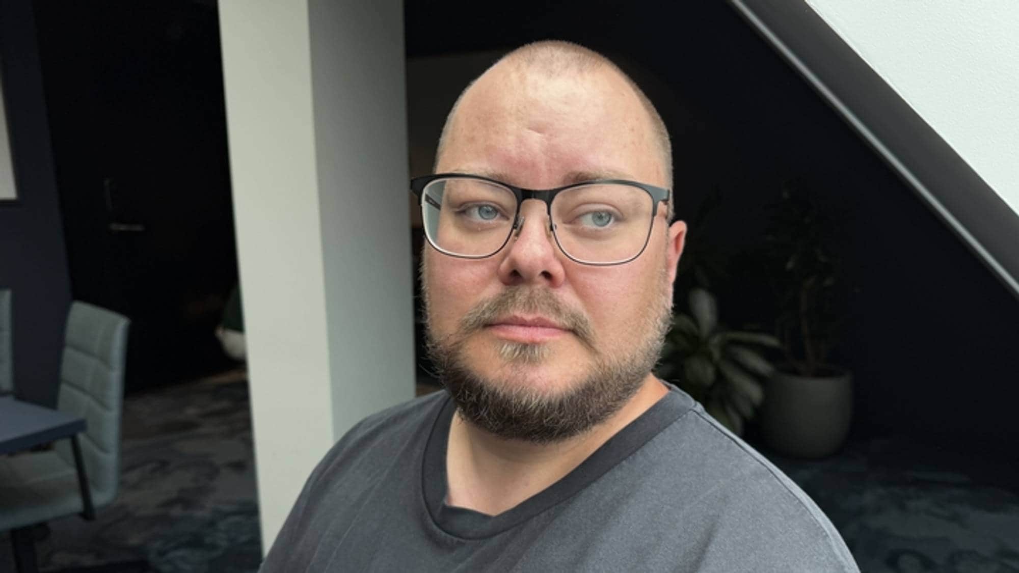 Mann med briller står i ved et vindu og ser ned til høyre. Han er ikledd en svart t-skjorte med trykk. 