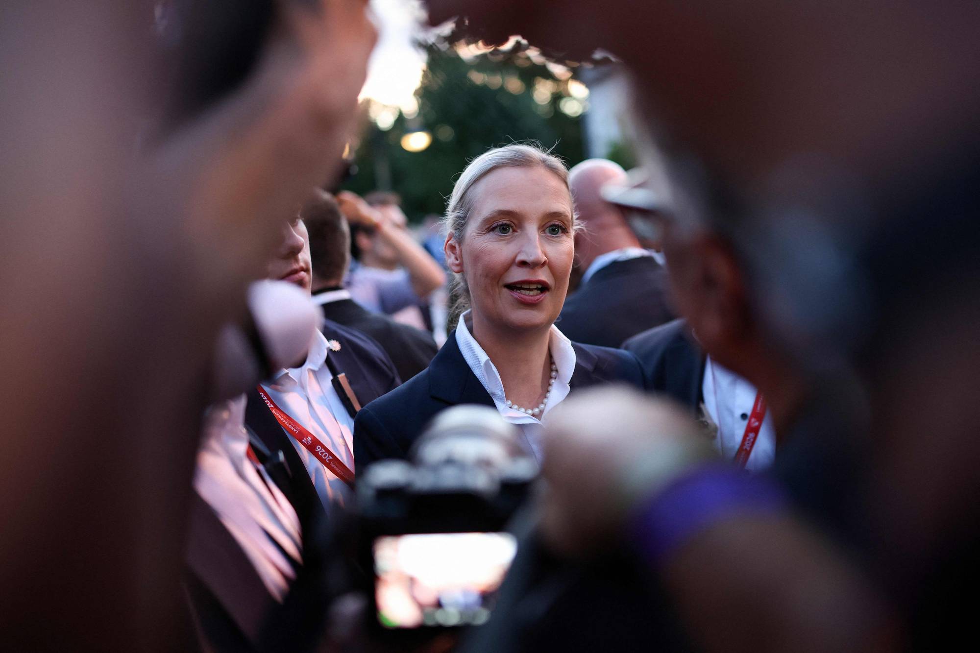 En kvinne med blondt hår i knute, dressjakke og hvit skjorte er omgitt av journalister og fotografer.