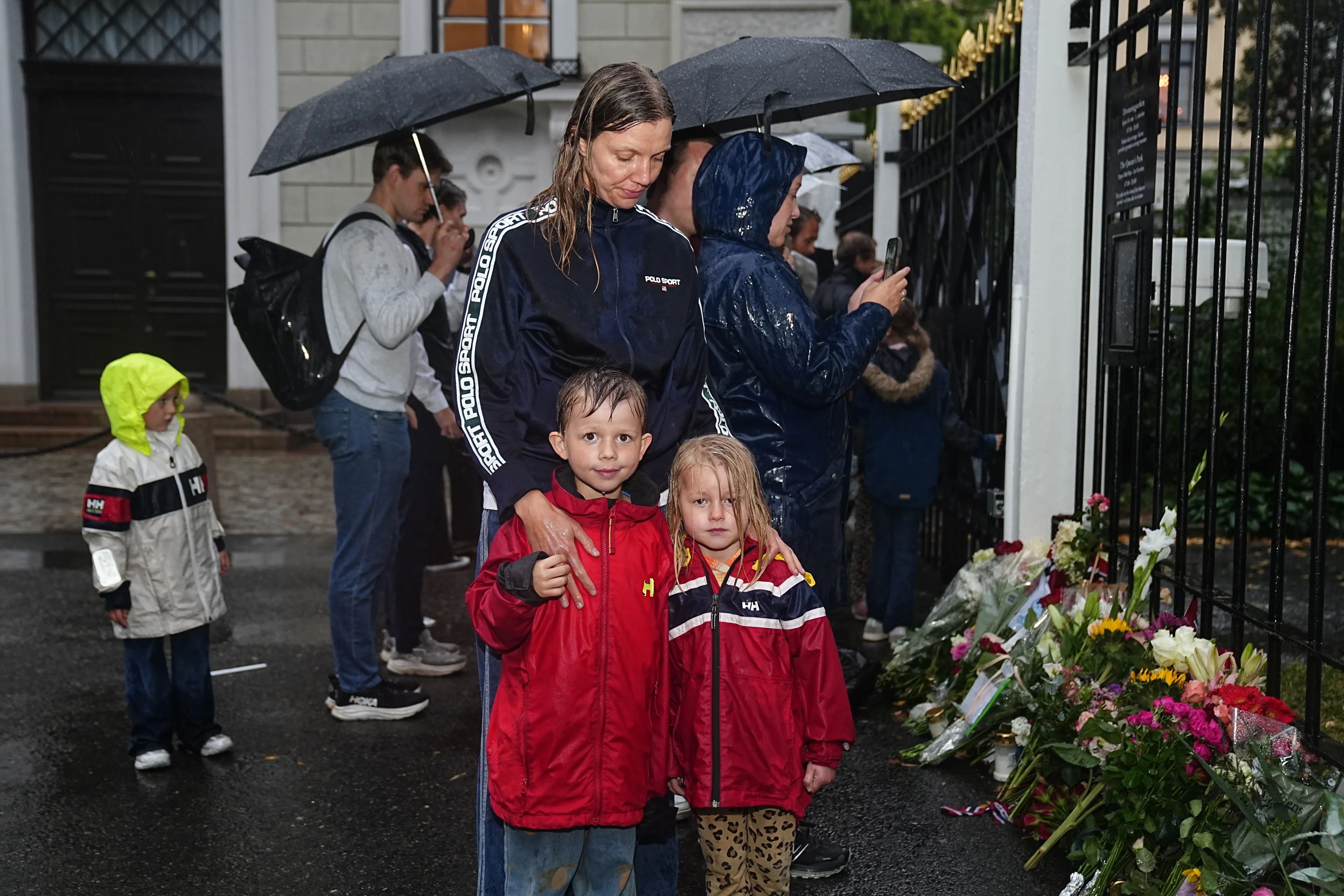 Folk legger ned blomster ved Slottets gjerde i regnvær. En mor står under en paraply sammen med sine to små barn. En liten gutt og jente står med vått hår i hver sin røde regnjakke.