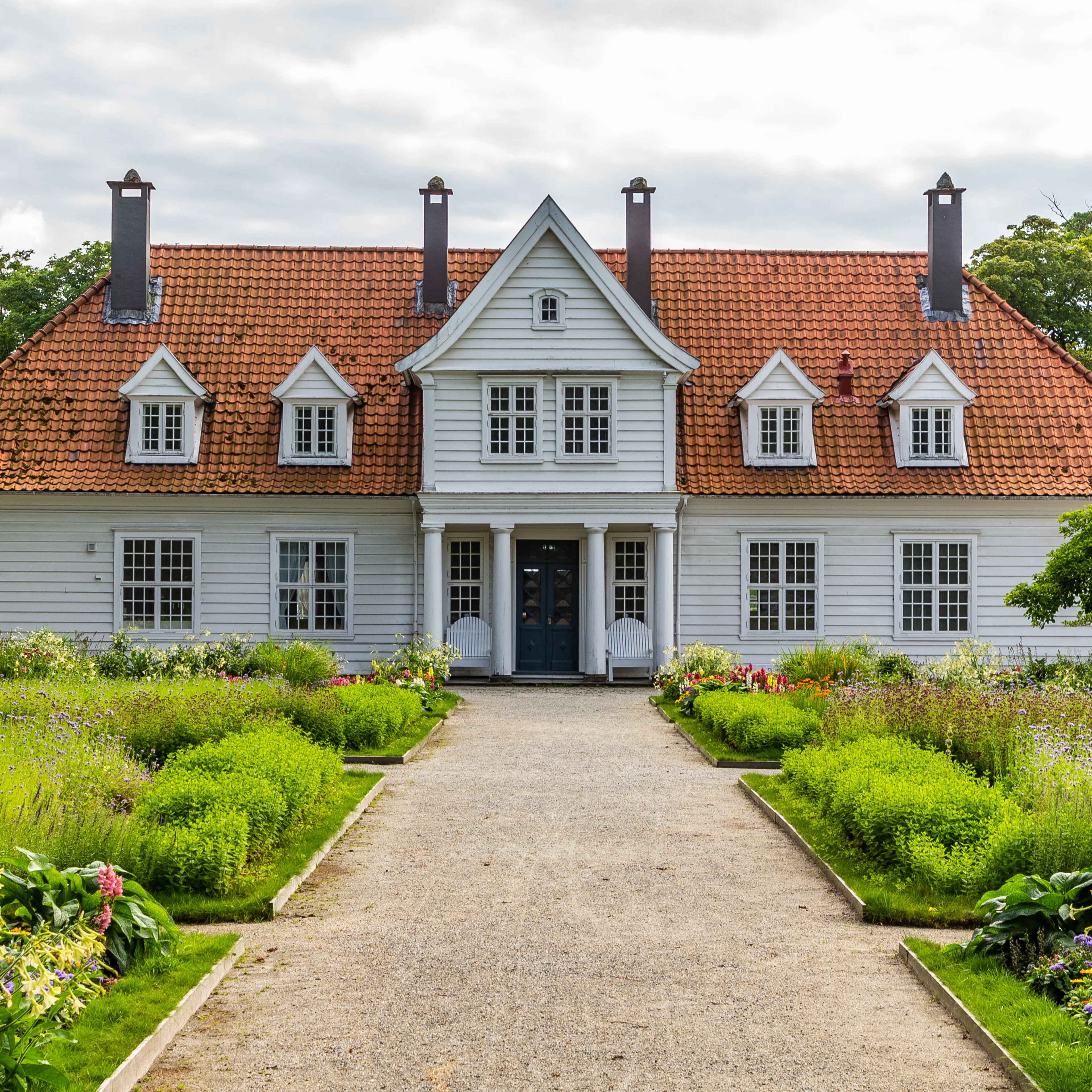 Hvit trebygning med rødt tegltak sett fra en hagegang med blomsterbed.