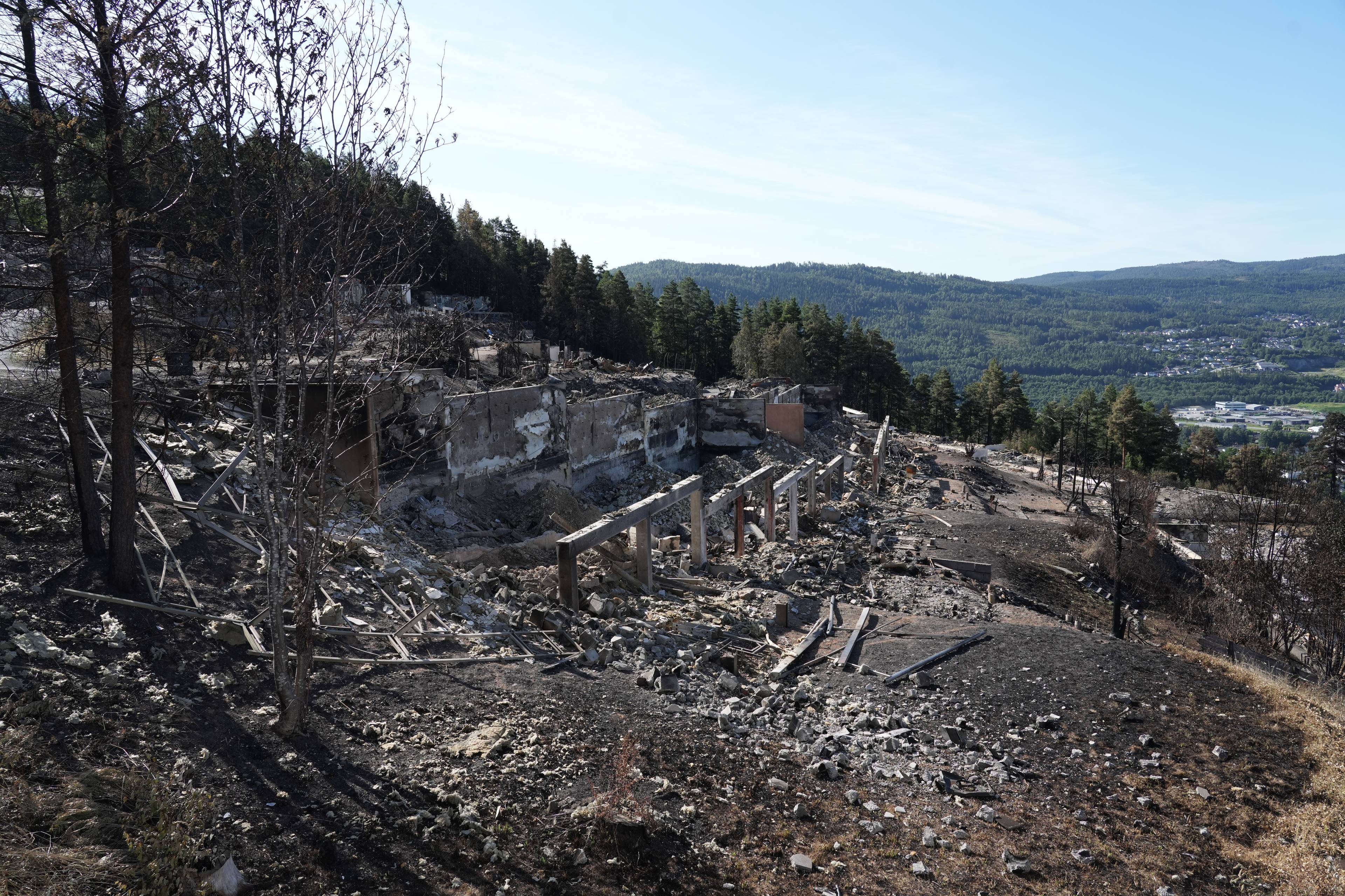 Ruiner etter en brann i en bygning i en skråning med skog og bebyggelse i bakgrunnen. (Bildebeskrivelsen er laget av en KI-tjeneste)