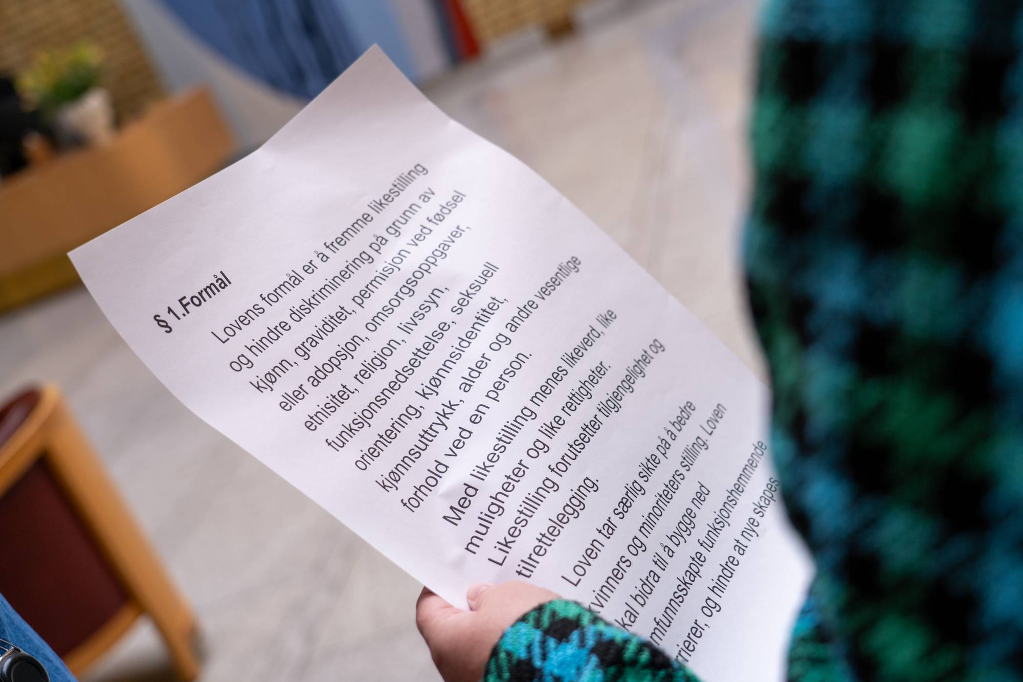 En person holder et papir med tekst.
Teksten på papiret er et utdrag fra en lov om likestilling.
Den beskriver formålet med loven og hva som menes med likestilling.
Papiret holdes opp foran en person med et grønt og blått mønstret plagg. (Bildebeskrivelsen er laget av en KI-tjeneste)