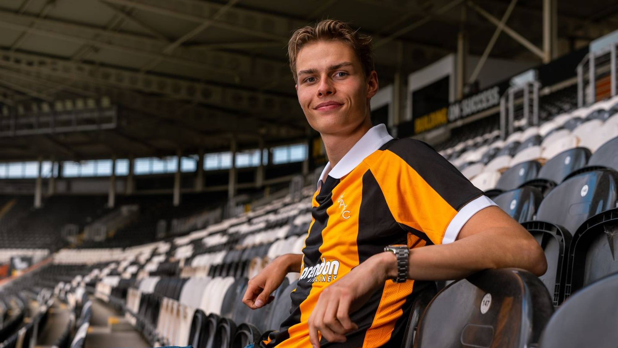 Ung mann i Hull City-drakt sitter på tribunene på et fotballstadion. (Bildebeskrivelsen er laget av en KI-tjeneste)