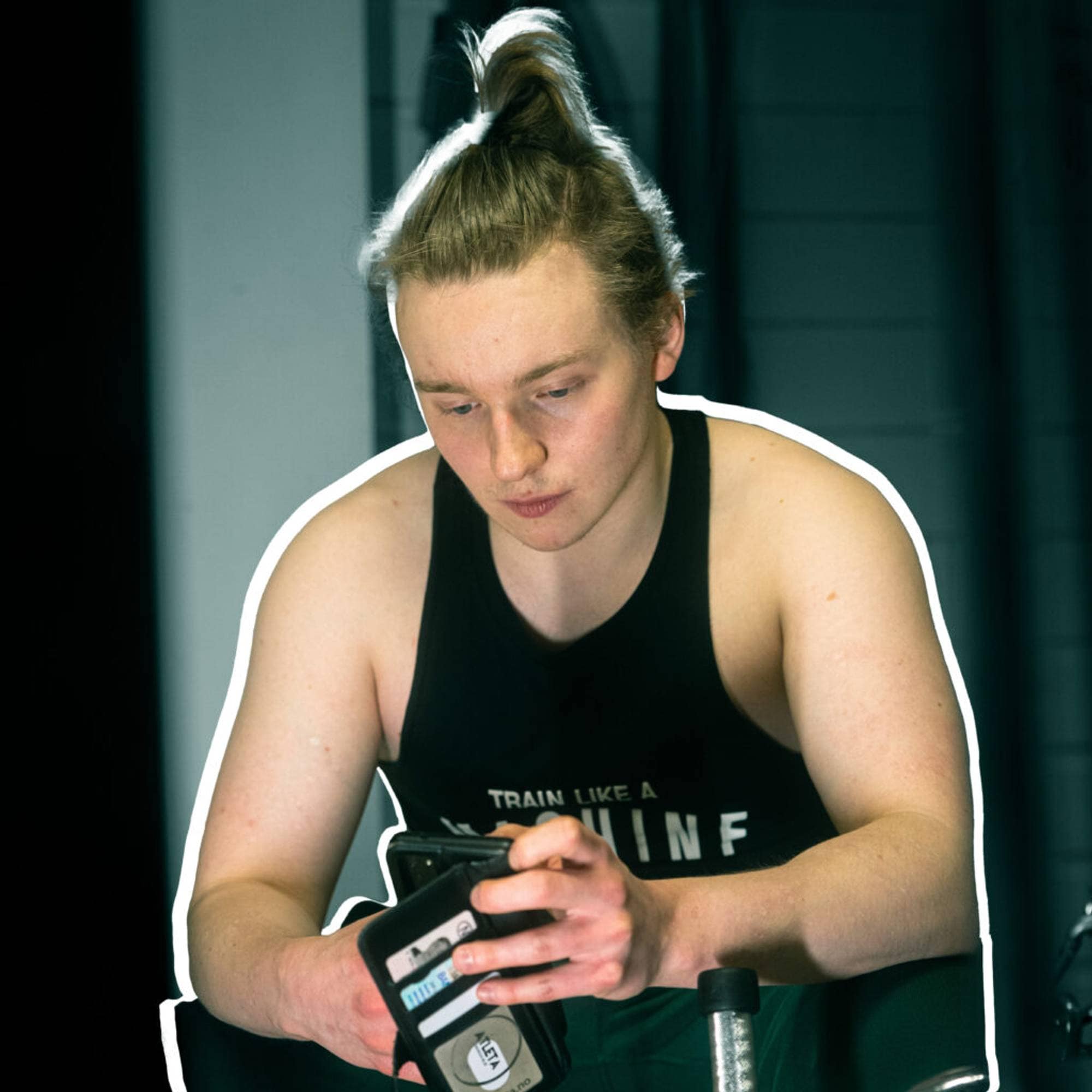 Bilde av Ian som scroller på telefonen. Han har på seg en singlet hvor det står "train like a machine" og grønne joggebukser. Håret er gyllenbrunt og i en dott på hodet. 