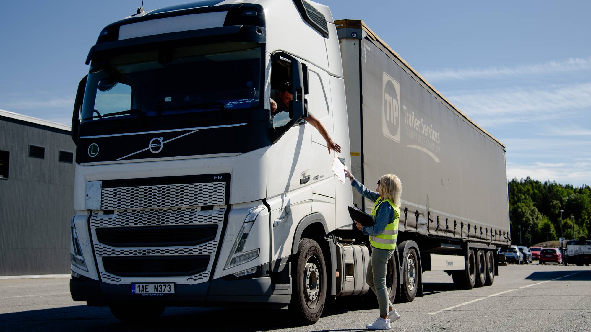 En kvinen i gul refleksvest står ved en lastebil og rekker noe inn vinduet til sjåføren. 