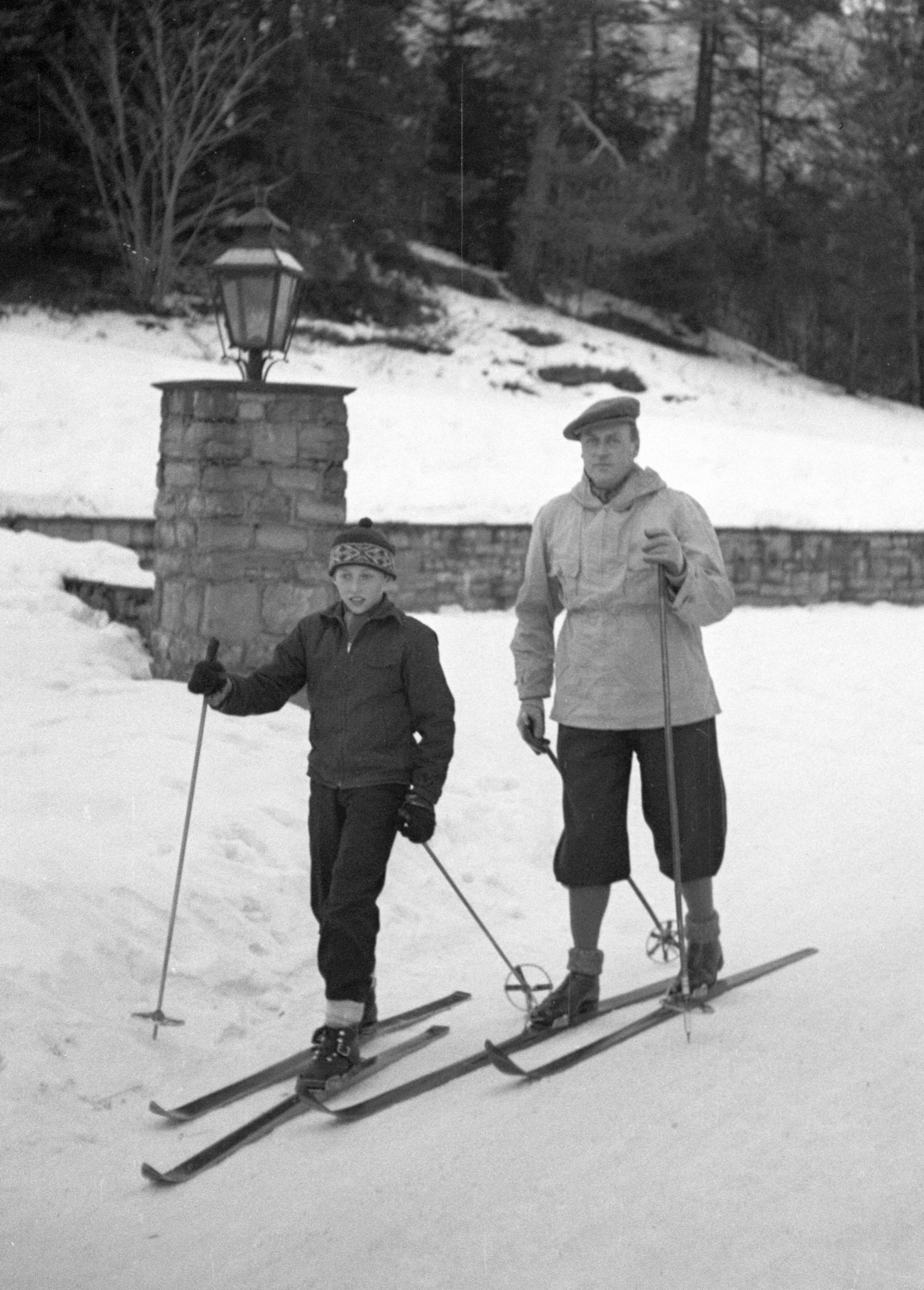 En gutt og en voksen mann står på ski i et snødekket landskap ved en lyktestolpe. Svart-hvitt foto. (Bildebeskrivelsen er laget av en KI-tjeneste)