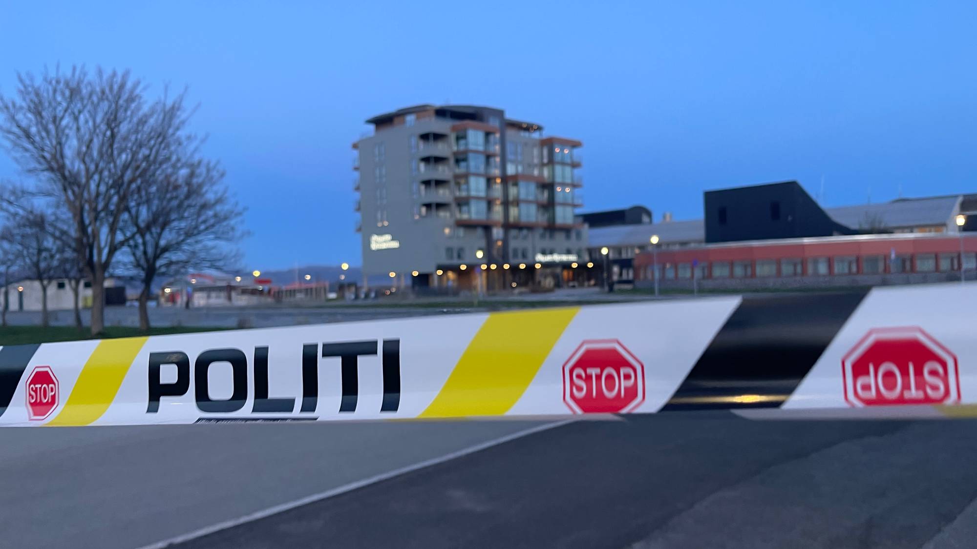 Politibånd sperrer av et område. Et moderne leilighetskompleks og andre bygninger er synlige i bakgrunnen. Skilt med "STOPP" er integrert i båndet. Deler av bildet er ute av fokus. (Bildebeskrivelsen er laget av en KI-tjeneste)