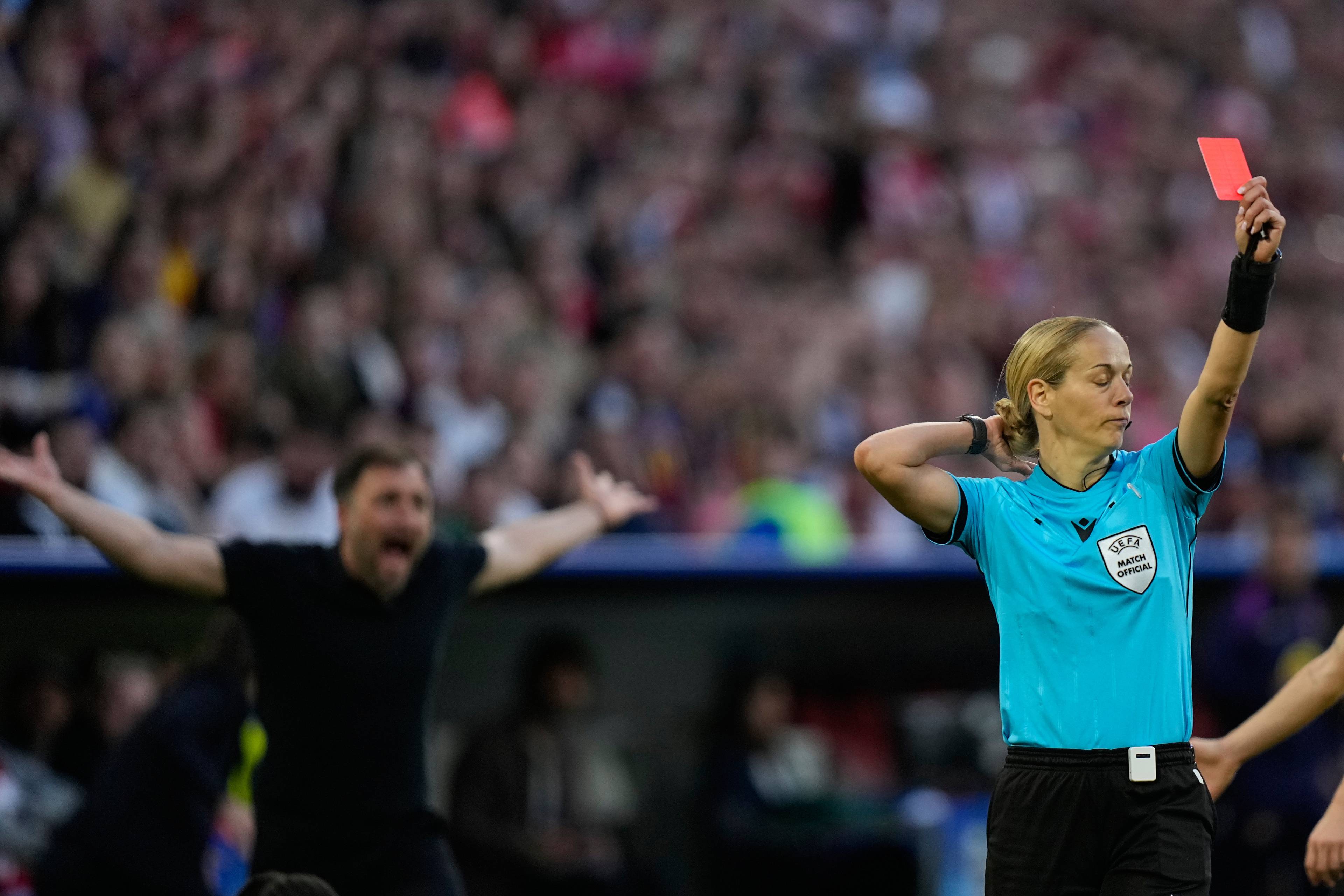 Dommeren holder opp et rødt kort under en fotballkamp. En mann med armene ut i luften ser ut til å protestere. Tilskuere sitter på tribunen i bakgrunnen. (Bildebeskrivelsen er laget av en KI-tjeneste)