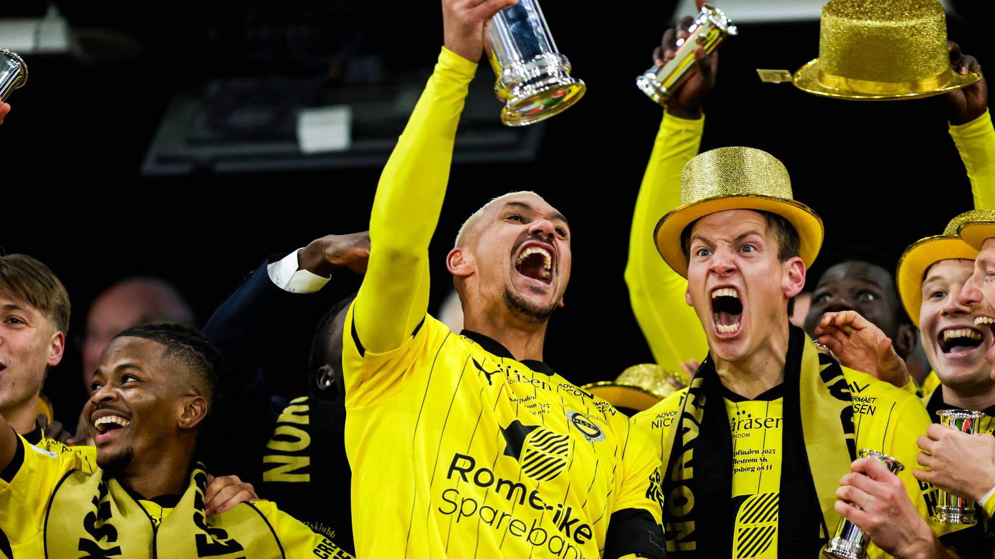 En gruppe mennesker feirer med trofeer under en seremoni. De har på seg gule trøyer og gule hatter. Mange av deltakerne smiler, mens noen holder opp pokaler. Omgivelsene tyder på en sportsbegivenhet med festlig stemning. (Bildebeskrivelsen er laget av en KI-tjeneste)