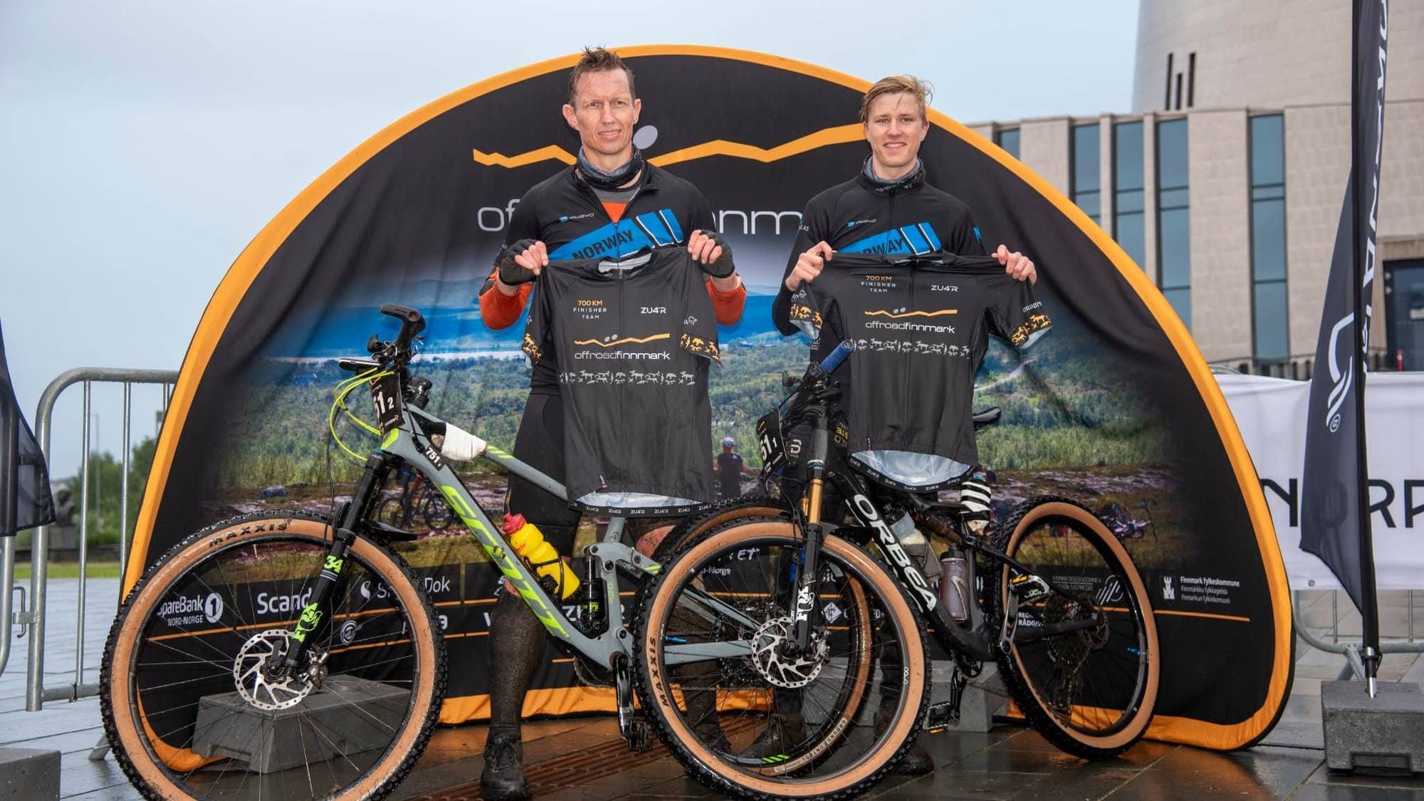 To syklister viser frem finisher-trøyer foran Offroad Finnmark-skilt og terrengsykler. (Bildebeskrivelsen er laget av en KI-tjeneste)