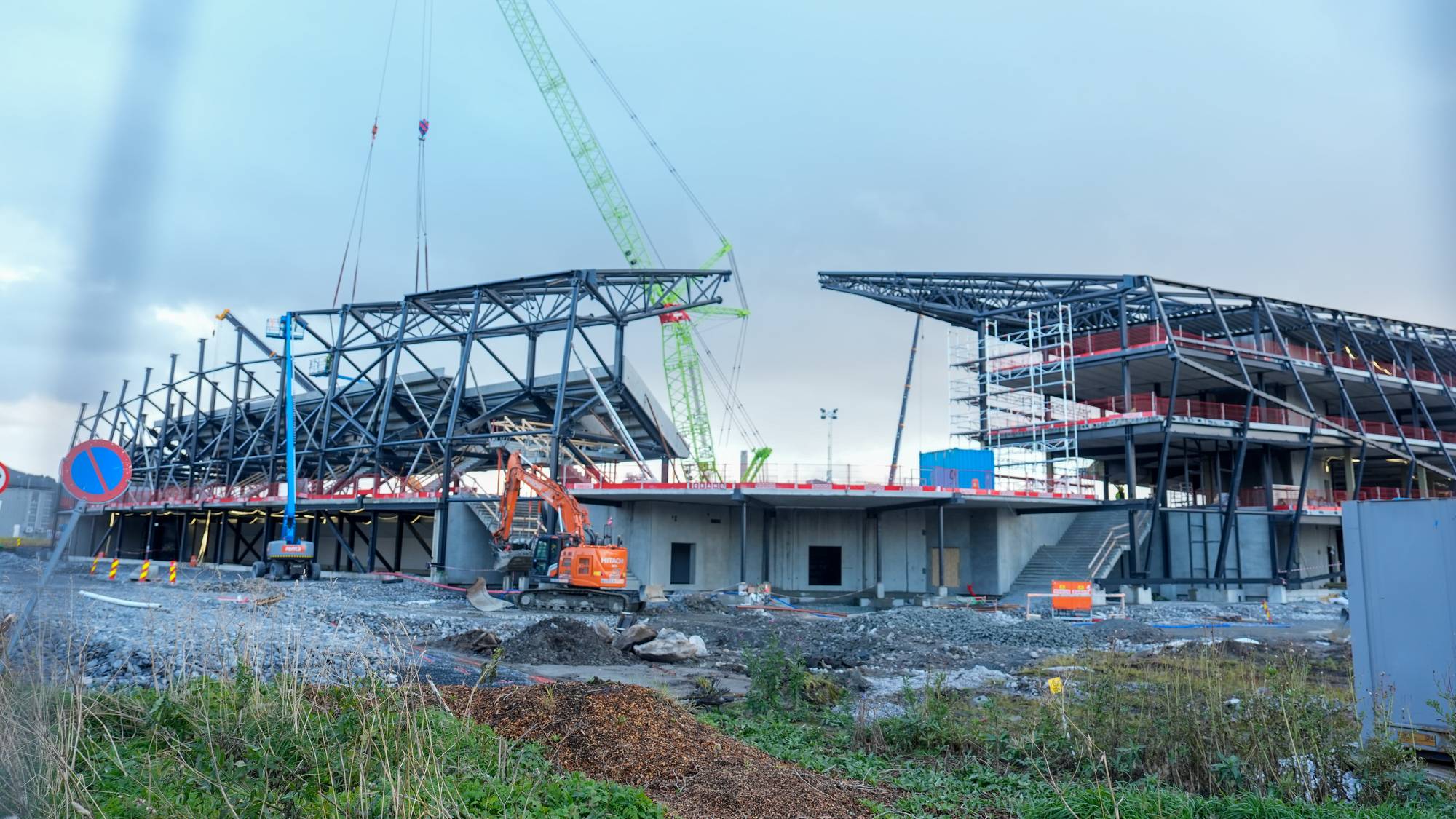 Byggeplass for idrettsstadion med stålkonstruksjoner, gravemaskin og stor kran under oppføring. (Bildebeskrivelsen er laget av en KI-tjeneste)