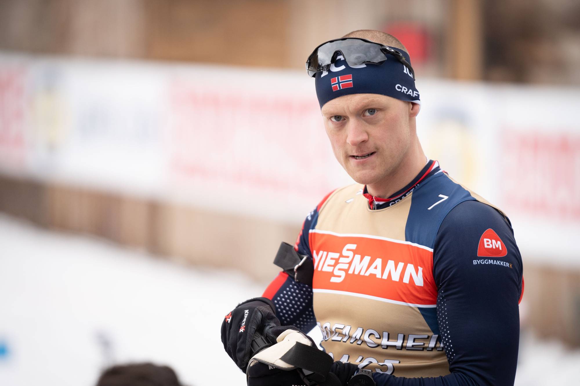 En idrettsutøver i langrennsklær. Han har på seg en blå lue med norsk flagg og solbriller på hodet. Han holder hanskene sine. Omgivelsene er snødekte og uklare. (Bildebeskrivelsen er laget av en KI-tjeneste)