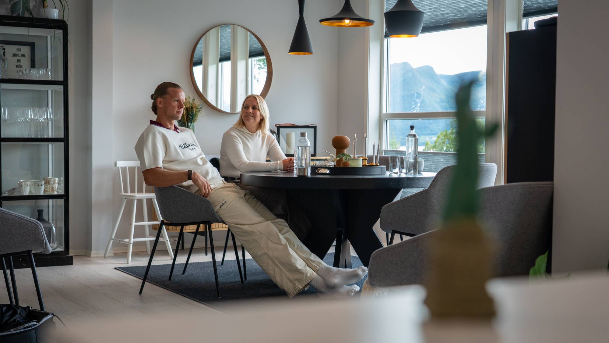 Mann og kvinne sitter ved et spisebord i en lys stue med utsikt mot fjell. (Bildebeskrivelsen er laget av en KI-tjeneste)