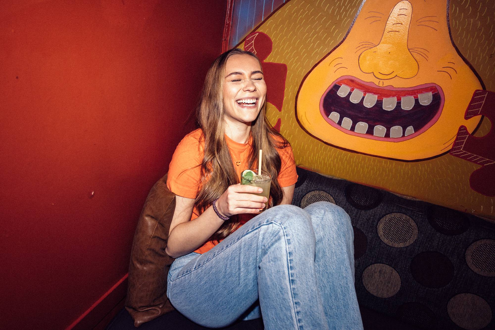Julie ler høyt mens hun sitter i en hjørnesofa. Hun har en alkoholfri drink i høyre hånd. Hun har på seg en jeans og en oransje t-skjorte. 