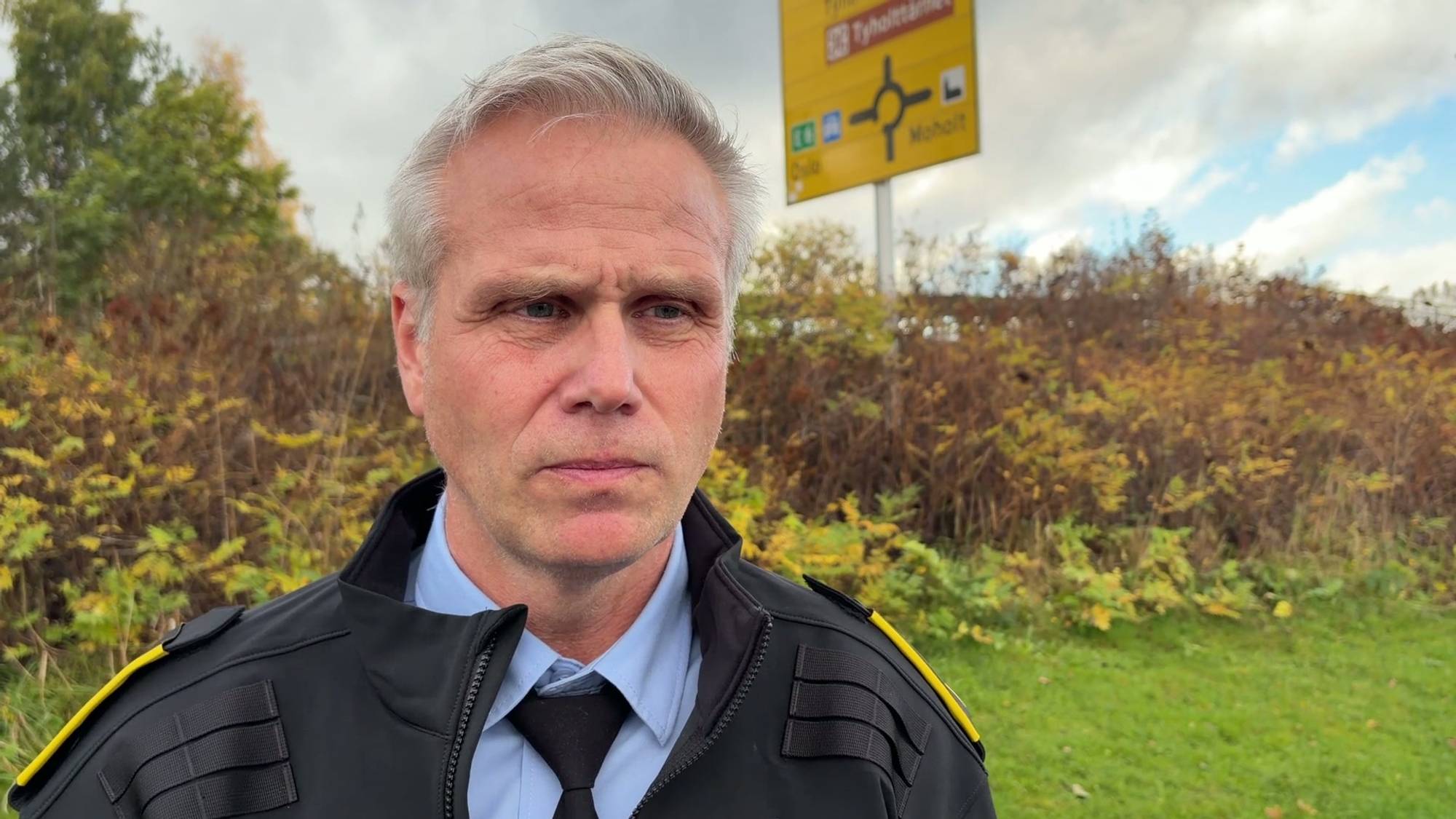 Politibetjent står utendørs ved et veiskilt med vegetasjon i bakgrunnen. (Bildebeskrivelsen er laget av en KI-tjeneste)