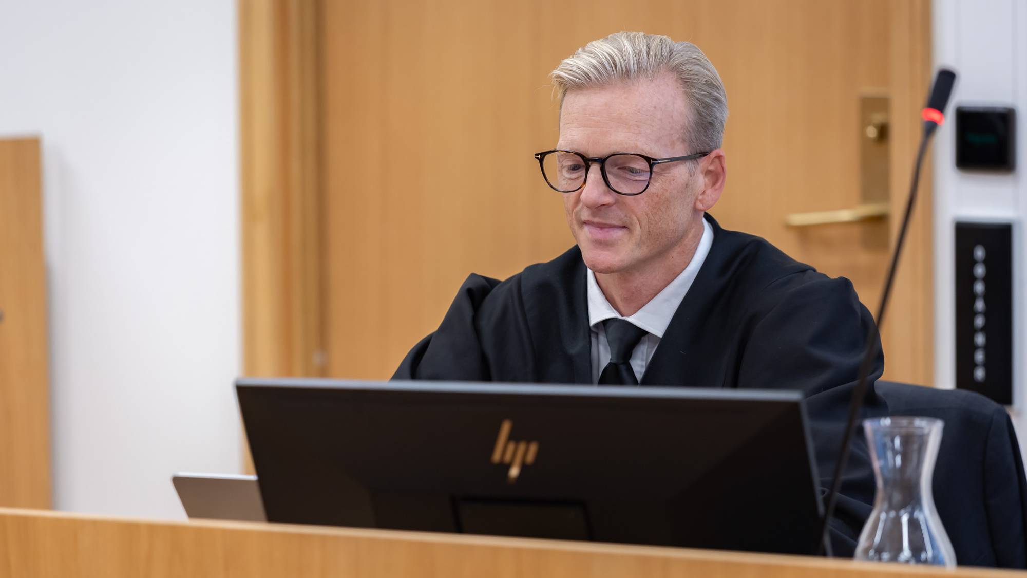 En mann iført svart kappe og svart slips sitter ved PC-en i en rettssal. Han kikker på en PC-skjerm og smiler forsiktig. Mannen har briller med mørk innfatning