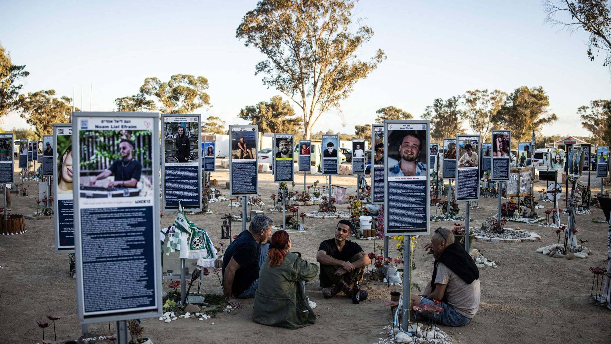 En familie sitter på bakken omringet av minnesmerker av de som ble drept 7.oktober av Hamas, på Nova-festivalen nær grensen til Gaza.
