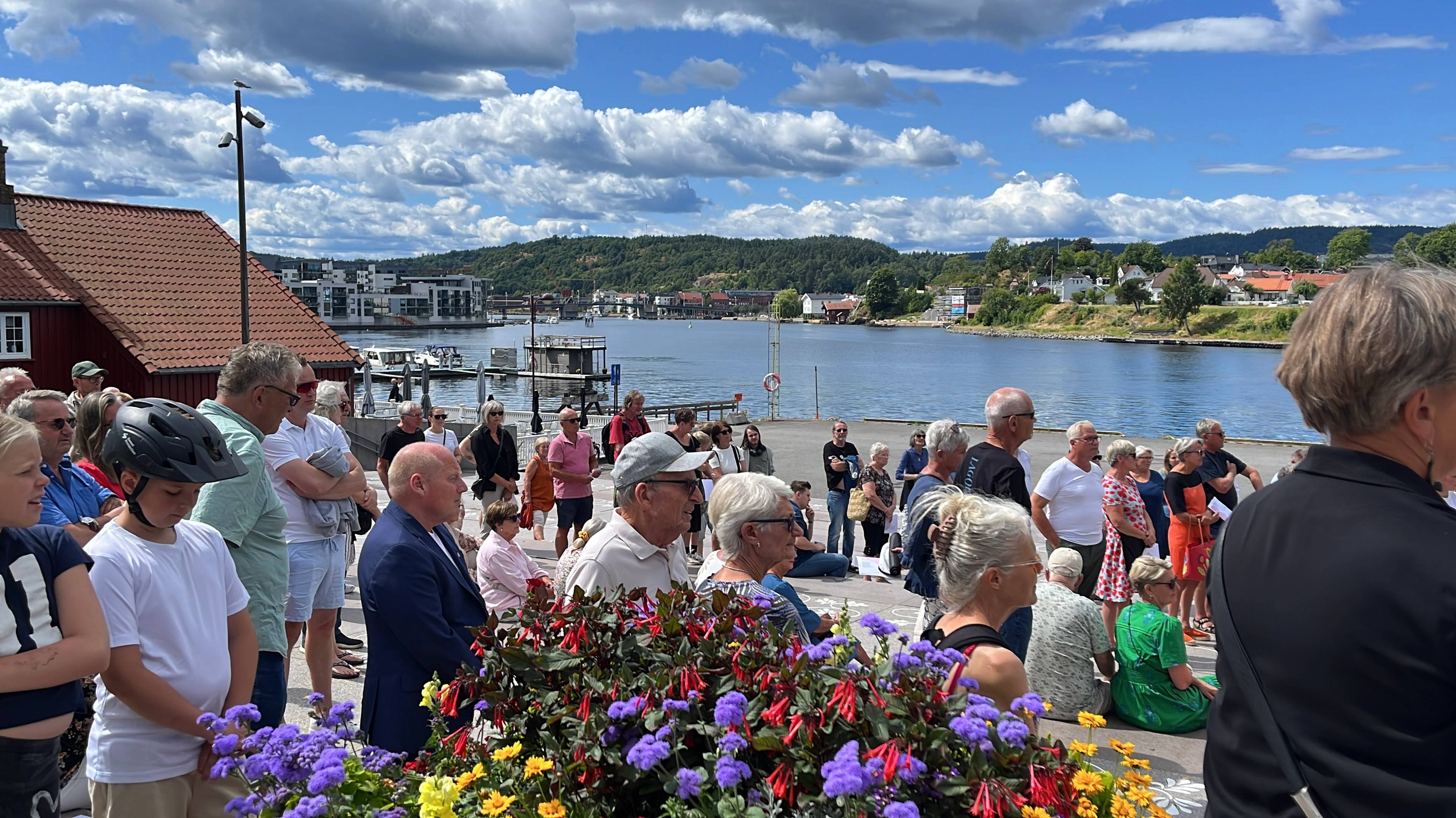 Folk er samlet utendørs ved sjøen i solfylt vær for å delta på et arrangement. (Bildebeskrivelsen er laget av en KI-tjeneste)