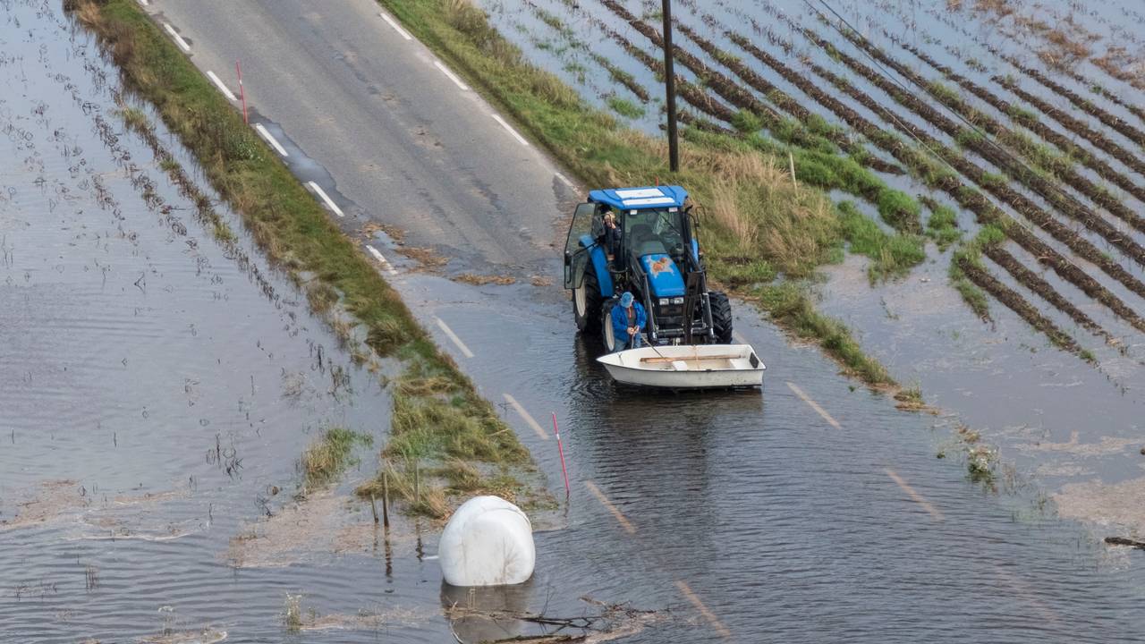 Vannet står høyt over veien. En traktor løfter en båt og en høyball i plast flyter i vannet.