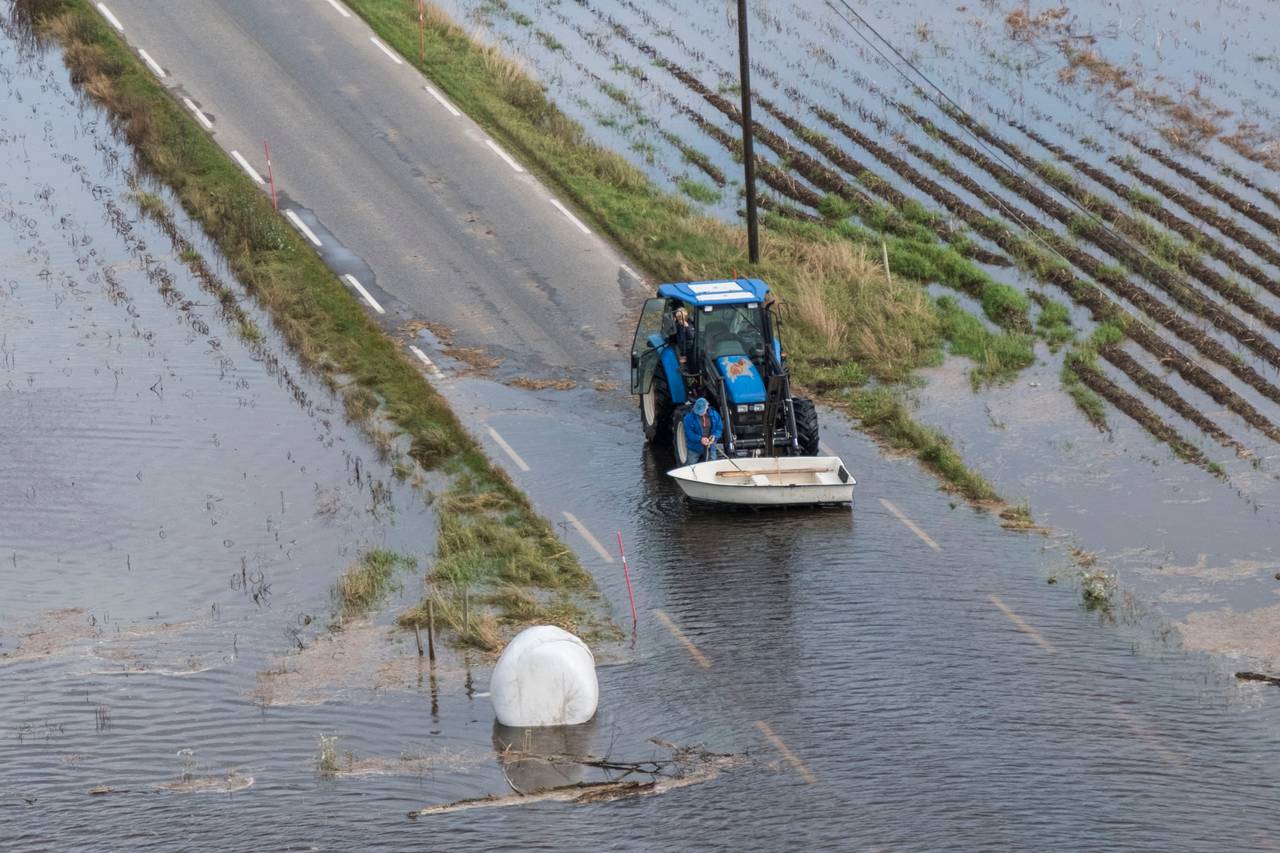 Vannet står høyt over veien. En traktor løfter en båt og en høyball i plast flyter i vannet.
