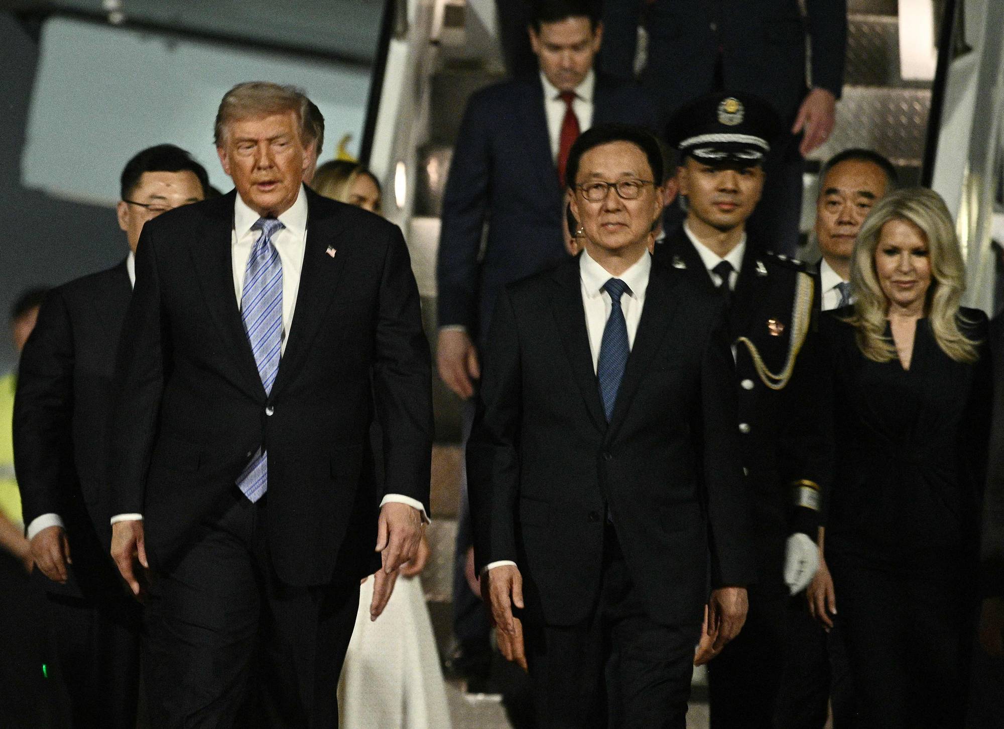 Donald Trump og en mann i mørk dress med briller går ned en trapp. En mann i militæruniform er bak dem. En kvinne i sort kjole går til høyre for den mannen i dress. (Bildebeskrivelsen er laget av en KI-tjeneste)