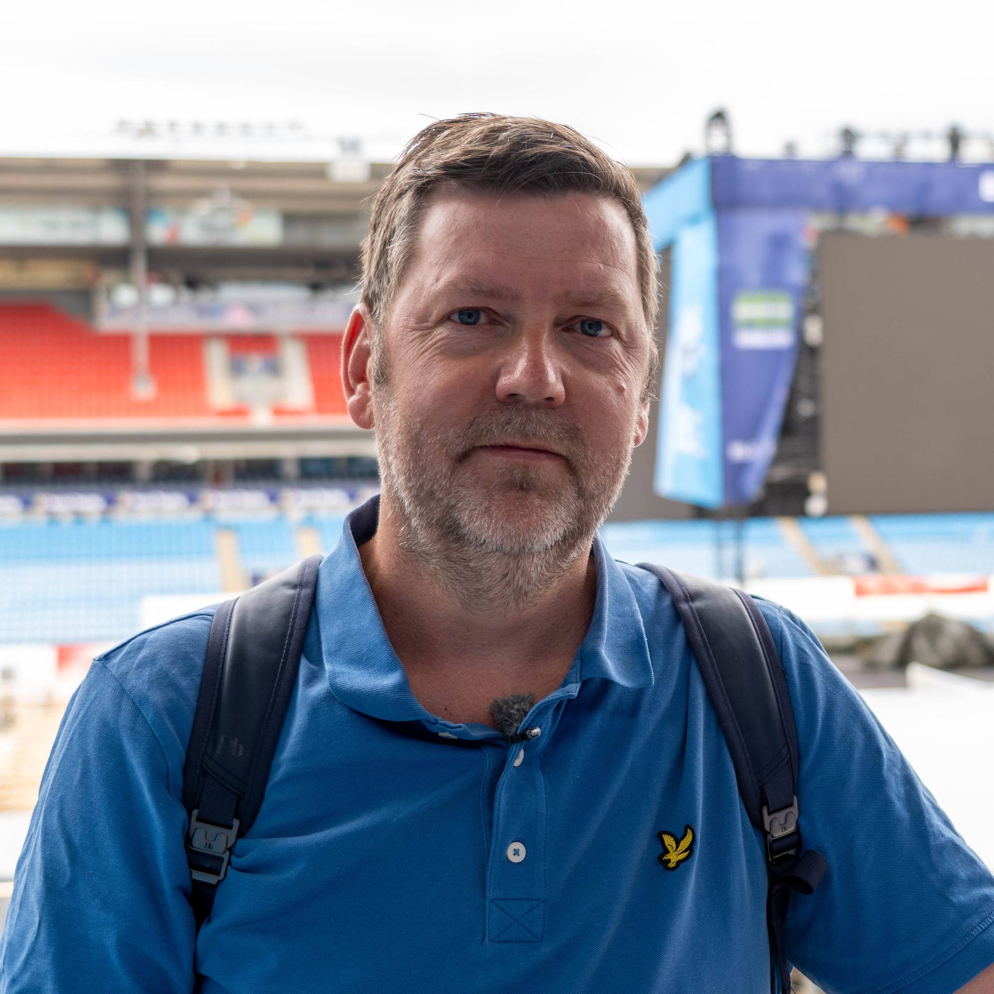 Mann i blå t-skjorte står foran en fotballstadion med en scene i bakgrunnen. (Bildebeskrivelsen er laget av en KI-tjeneste)