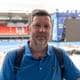 Foto: Roy Pettersen / NRK Mann i blå t-skjorte står foran en fotballstadion med en scene i bakgrunnen. (Bildebeskrivelsen er laget av en KI-tjeneste)