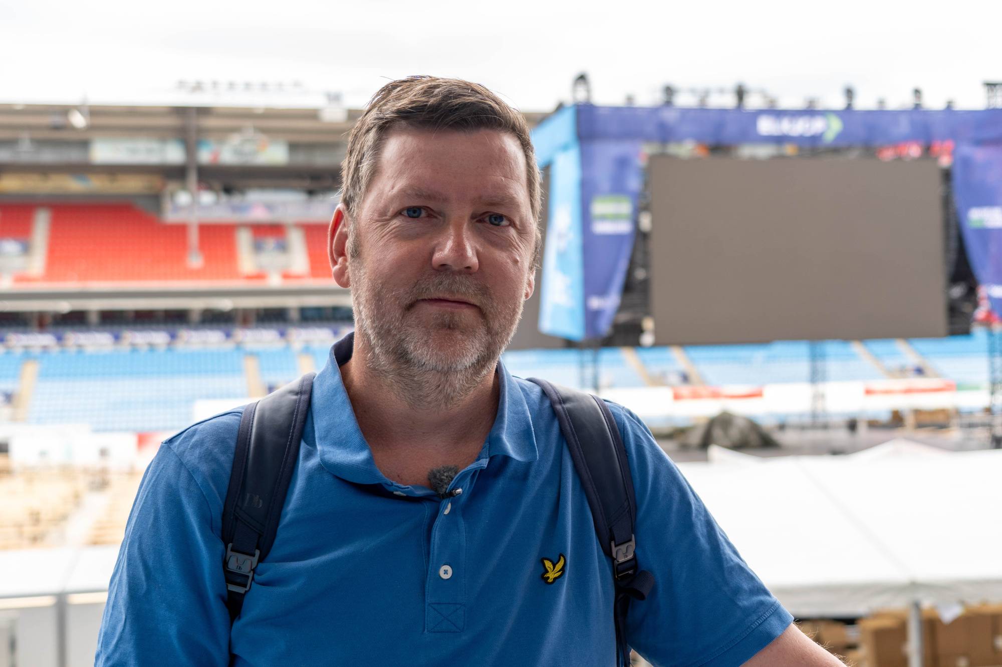 Mann i blå t-skjorte står foran en fotballstadion med en scene i bakgrunnen. (Bildebeskrivelsen er laget av en KI-tjeneste)
