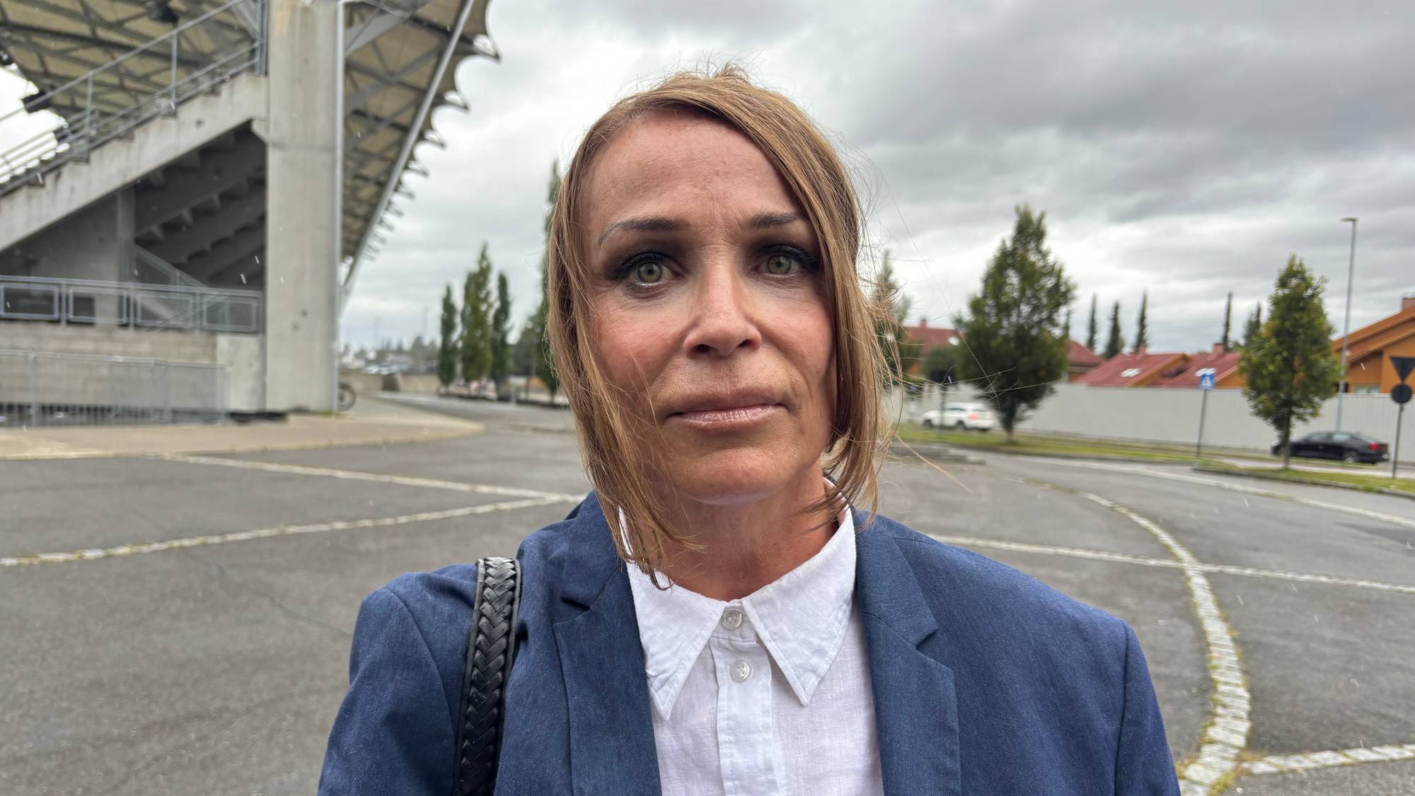 Kvinne i blå dressjakke står utendørs ved en stadionbygning. (Bildebeskrivelsen er laget av en KI-tjeneste)