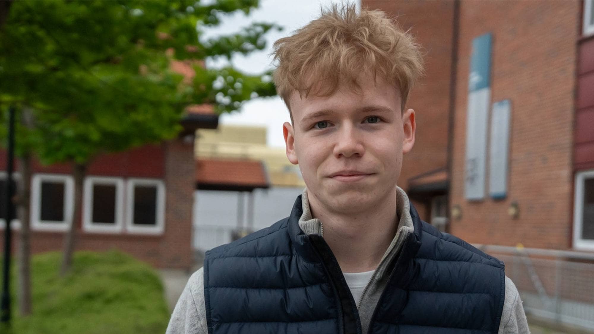 En gutt med blondt rotete hår står foran en skolebygning og ser rett i kamera.