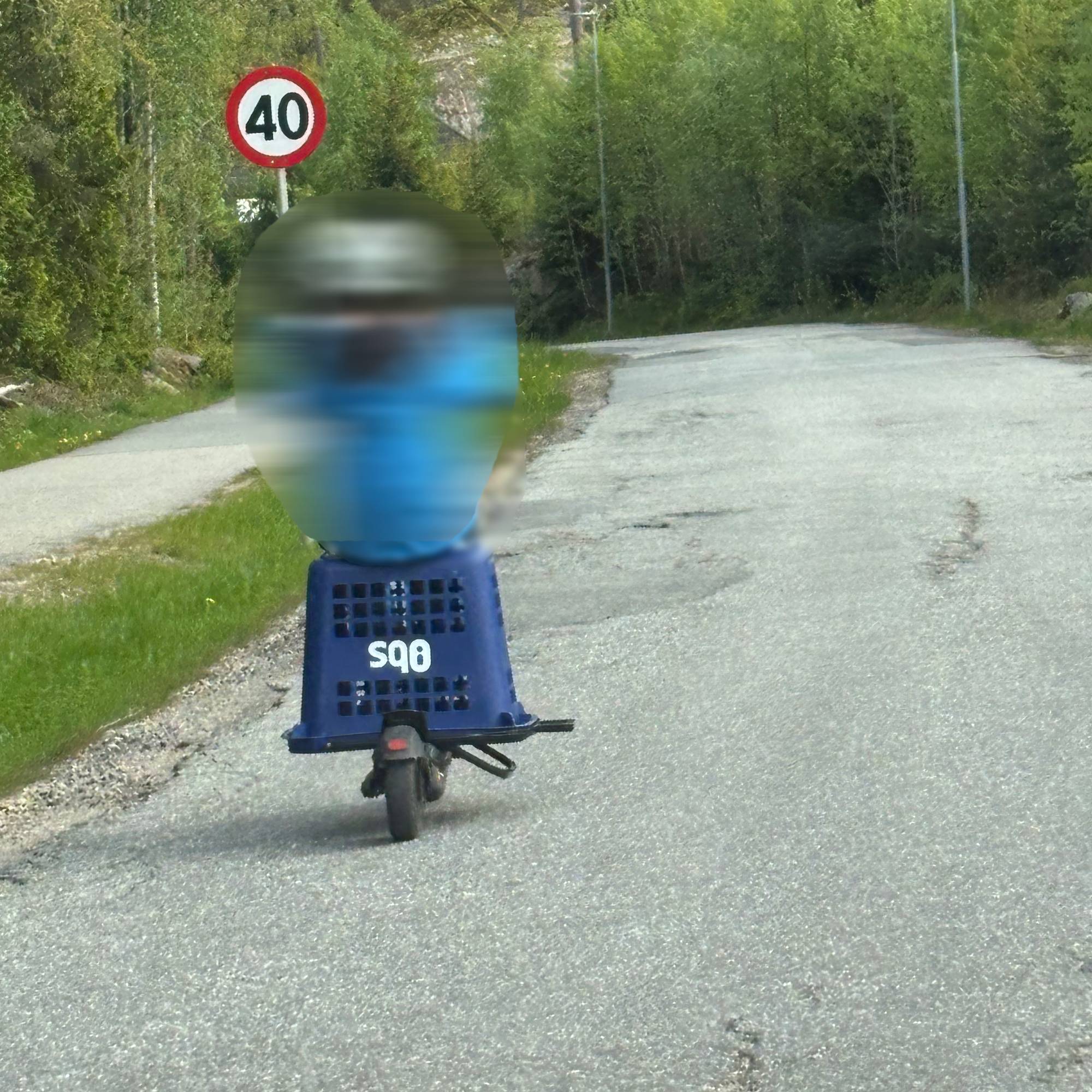 Et barn sitter på en handlekurv snudd opp ned og plassert på en elsparkesykkel. Barnet kjører nedover en vei.