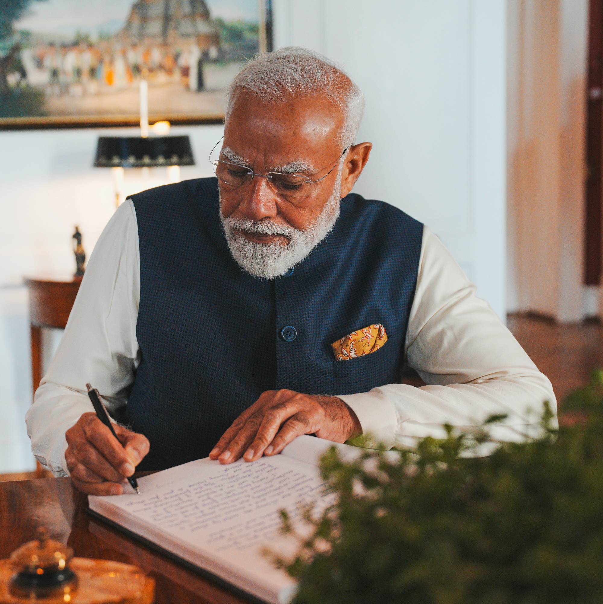 Indias statsminister Narendra Modi sitter ved et skrivebord og skriver i en bok. Han er iført en hvit skjorte og en mørkeblå vest.