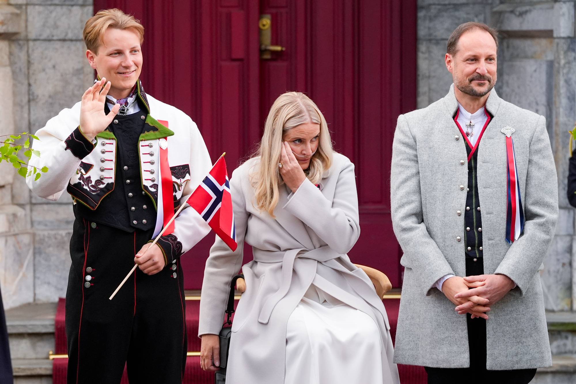 Kronprins Haakon, kronprinsesse Mette-Marit og prins Sverre Magnus vinker til barnetoget på Skaugum på 17. mai 2026. Kronprinsessen sitter på en stol og har nesekateter.