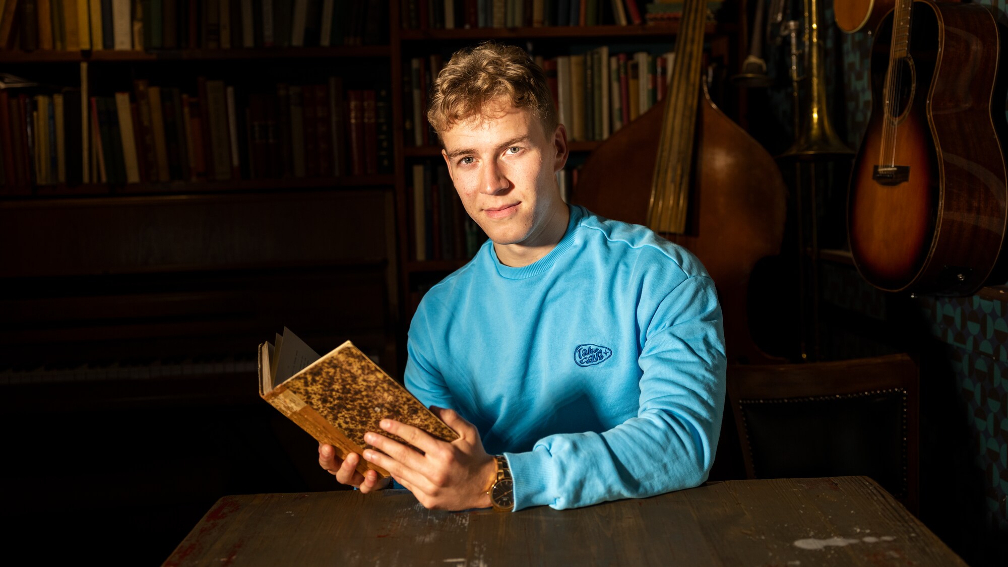 Joakim ser i kamera mens han holder i en bok. Han har på seg en blå sweatshirt. Han har lyse krøller og blå øyne. Han sitter foran en bokhylle.