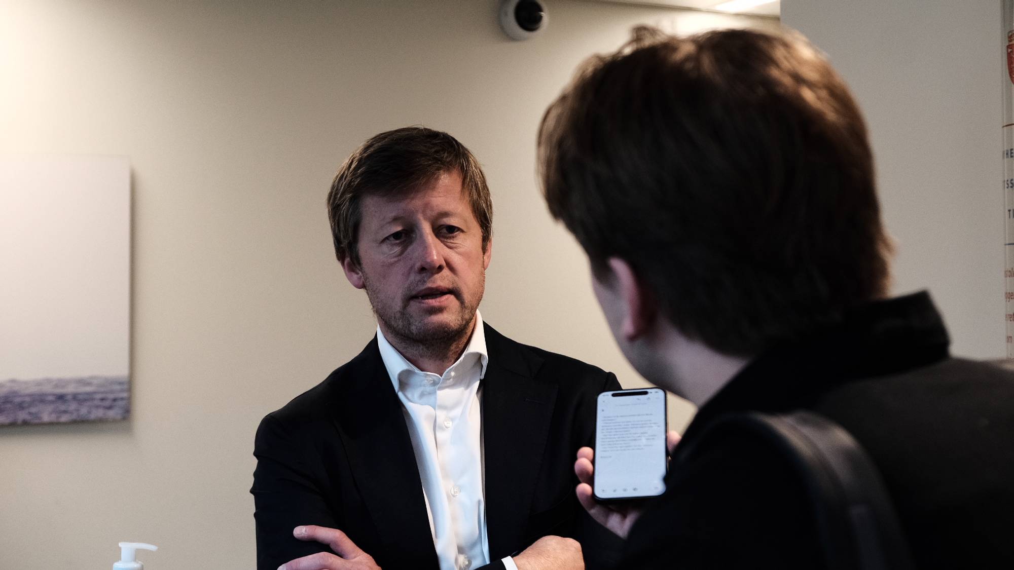 En mann i dress snakker med en annen person som holder en mobiltelefon foran ham. (Bildebeskrivelsen er laget av en KI-tjeneste)