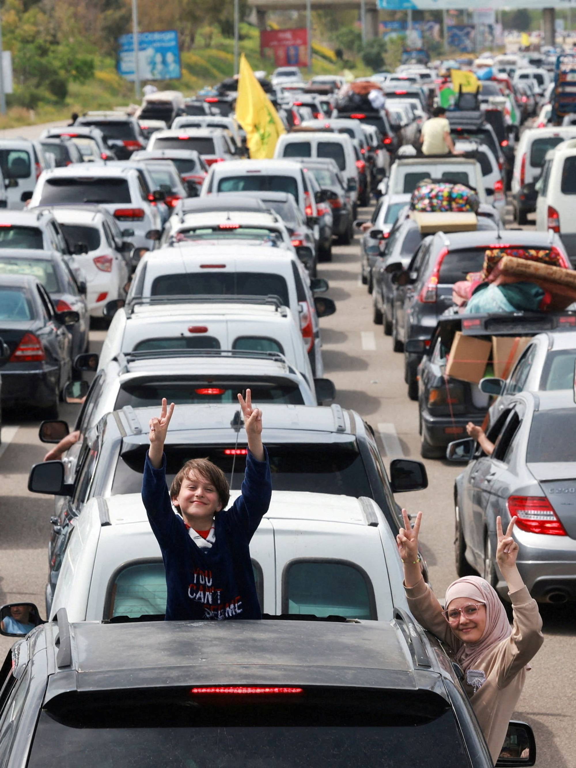 En gutt og en kvinne vinker fra biler i en trafikkork. Begge viser fredstegnet med fingrene. Biler står tett i tett på en vei, og noen har bagasje på taket. En gul flagg vaier i vinden fra en bil. (Bildebeskrivelsen er laget av en KI-tjeneste)